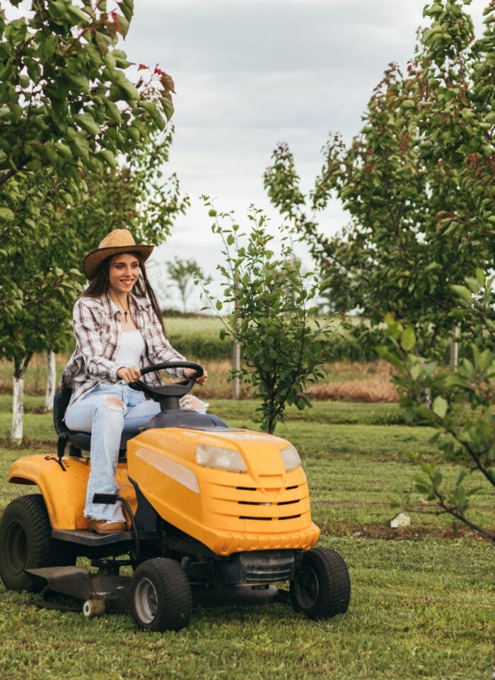 A woman driving a yellow lawn mower in an orchard.