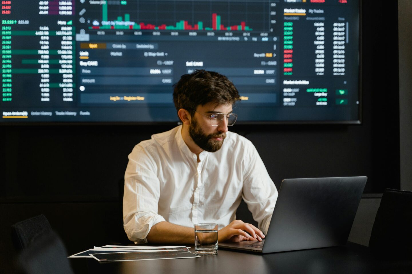 Traditional stockbroker working on a laptop in front of a large screen displaying live stock market data.