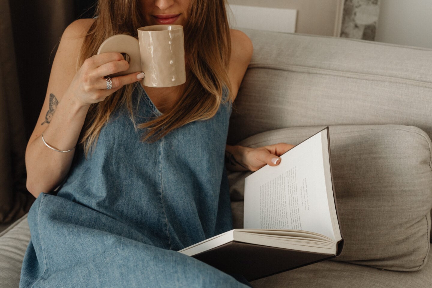 Woman sitting on a sofa reading a book while holding a mug, creating a calm and cozy self-care moment at home.