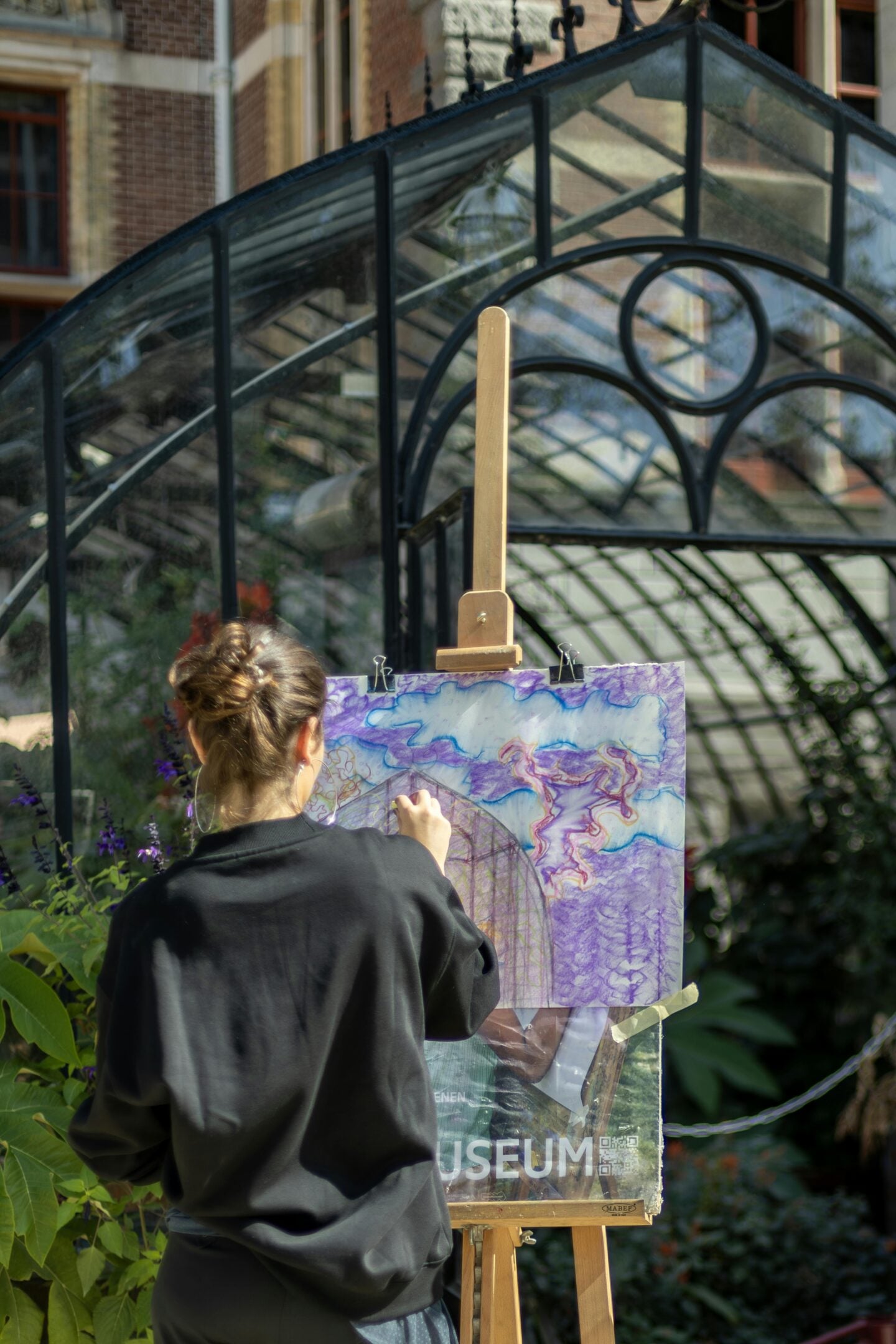 Artist painting on a canvas outdoors on a wooden easel, with a glass greenhouse structure in the background.