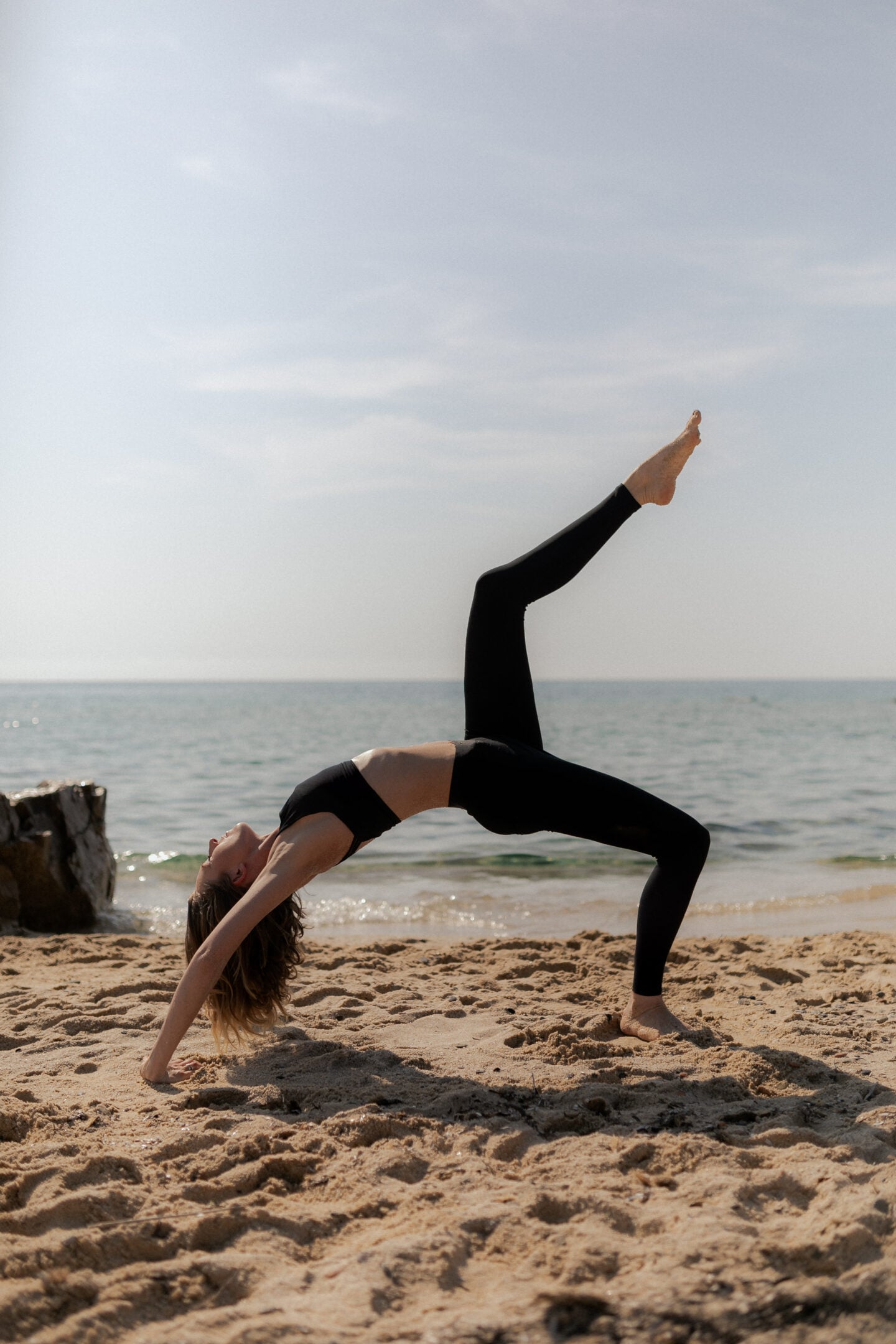 Person practicing yoga on a sandy beach, holding a backbend pose with the sea and horizon in the background.