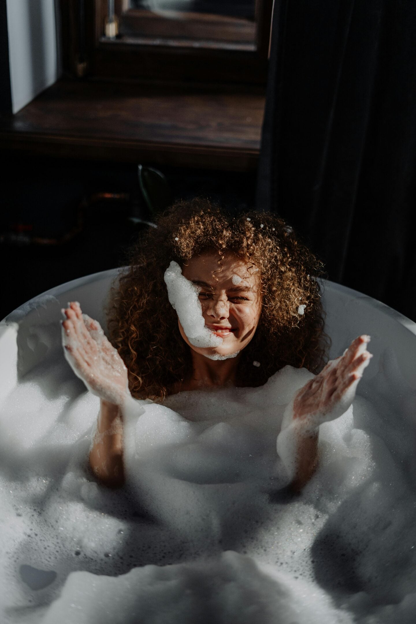 Person relaxing in a bathtub filled with foam, smiling while splashing water in a softly lit bathroom.