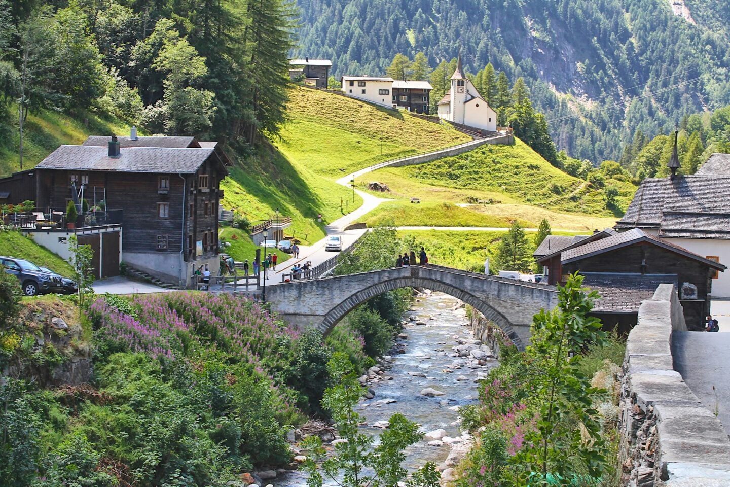 A picturesque Swiss mountain village with traditional wooden houses, a small stone arch bridge over a clear stream, a hillside church, and lush green alpine slopes in Binntal Nature Park, Wallis, Switzerland.
