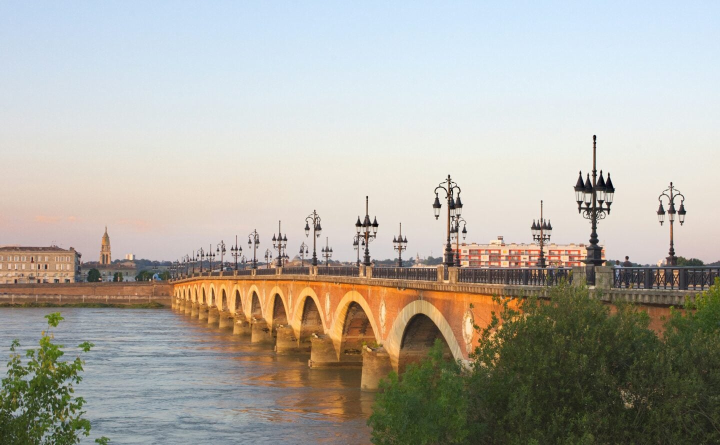 The Pont de Pierre bridge in Bordeaux at sunset, with its series of arches and ornate street lamps stretching across the Garonne River.