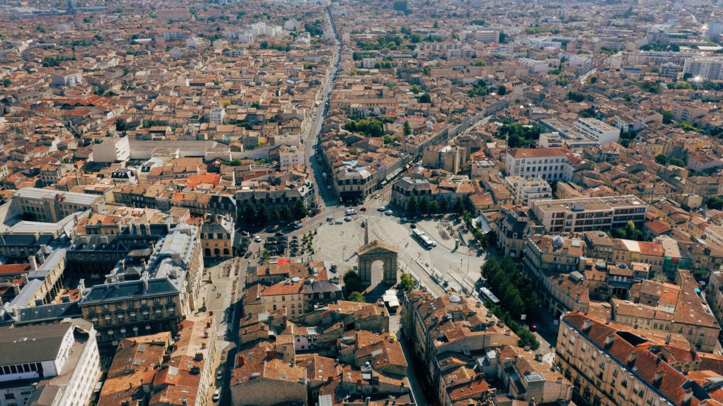 A panoramic aerial view of Bordeaux’s historic centre, showing terracotta rooftops, narrow streets, and the large square surrounding Porte de Bourgogne.