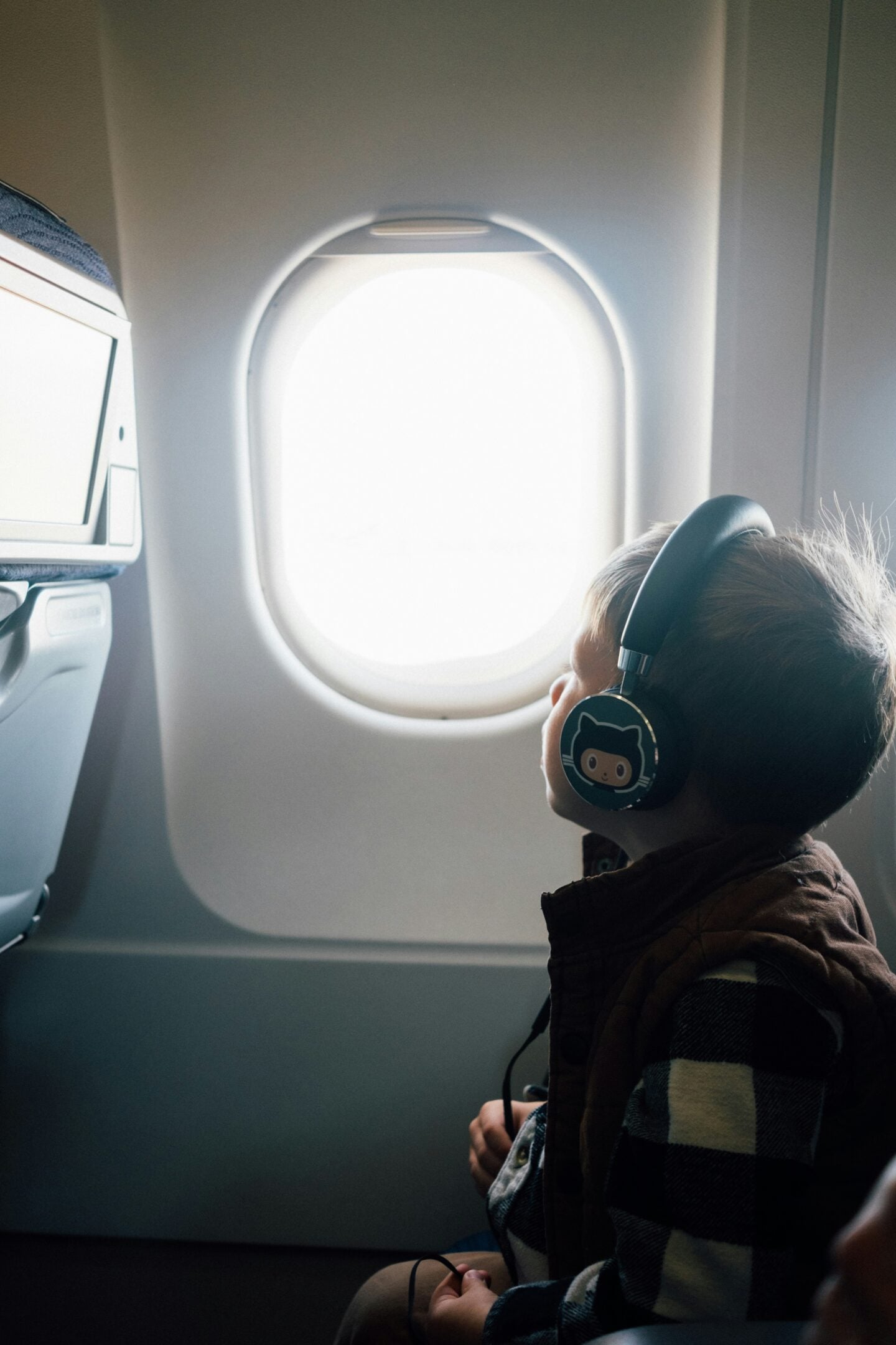 A young child wearing headphones looks out an airplane window during a flight, seated in the passenger cabin.