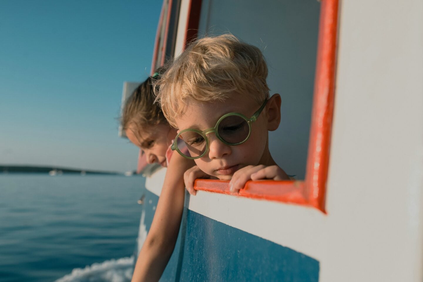 Two young children lean over the side of a boat, looking down at the water as the sun sets, with the boy wearing round green glasses.