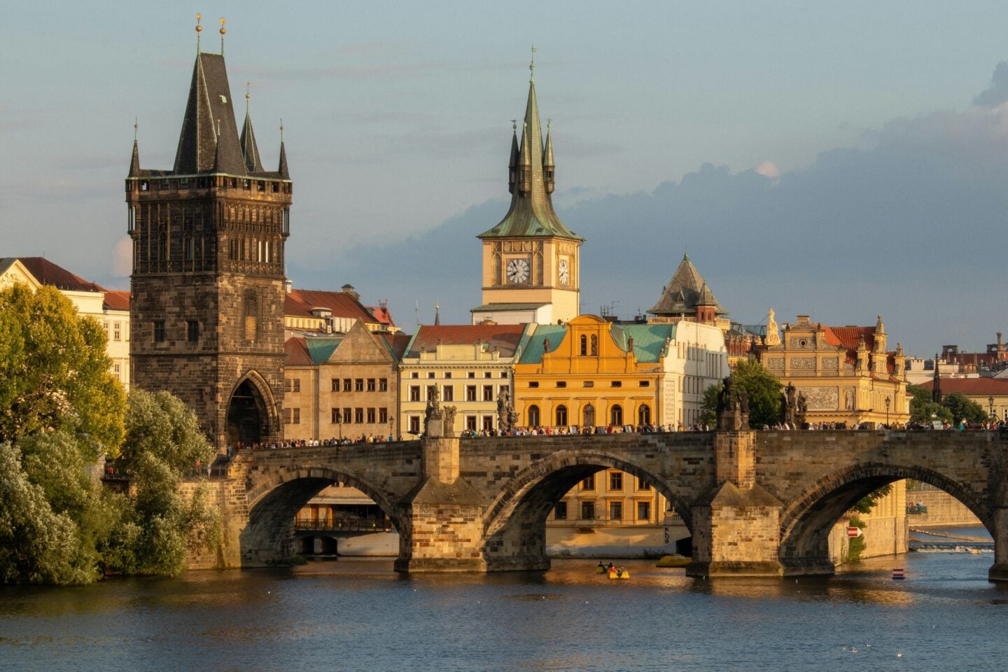 The Charles Bridge in Prague, Czechia, photographed at sunset with crowds walking across the historic stone arches and the Old Town Bridge Tower rising behind it.