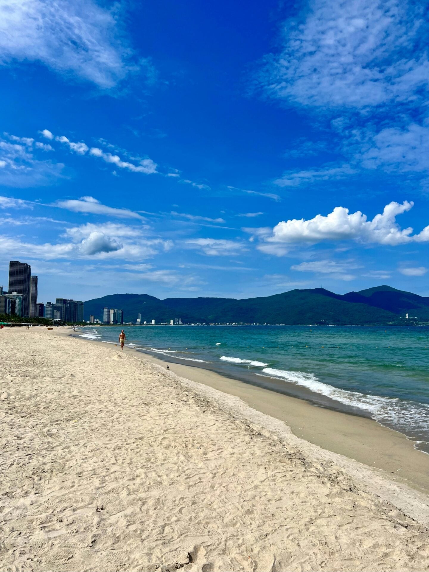 Sandy beach and turquoise sea in Da Nang, Vietnam, with the city skyline and mountains visible in the distance.