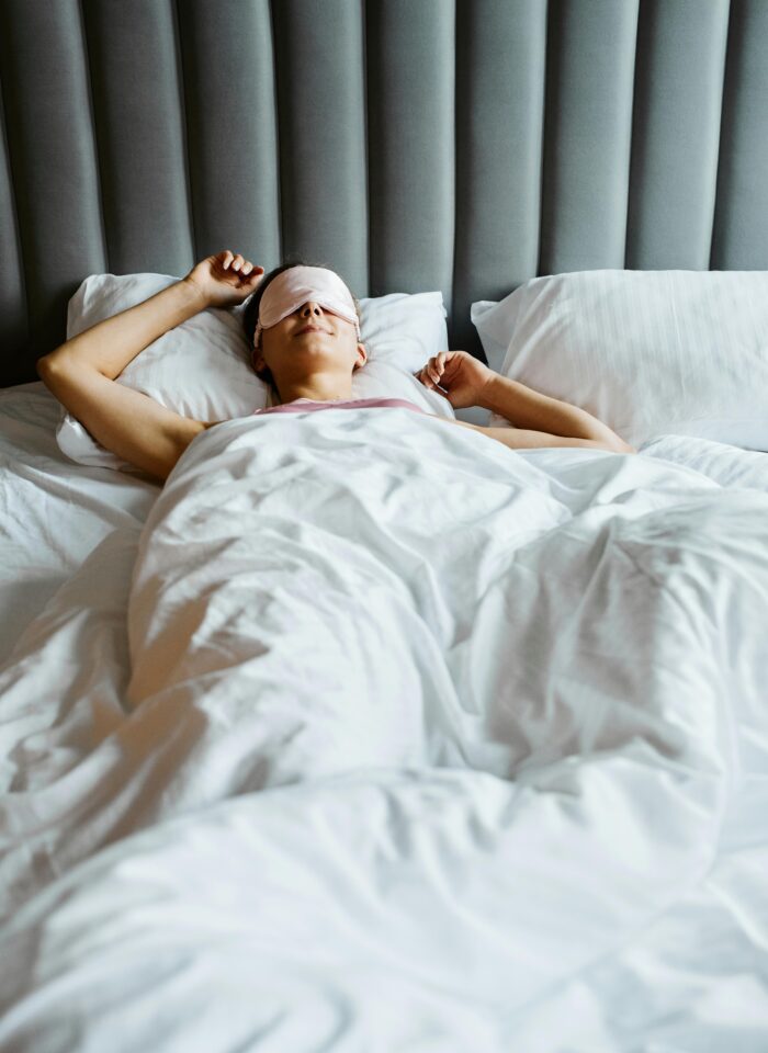A woman lies in bed under white blankets, wearing a pink sleep mask and resting comfortably against a tall, padded grey headboard.