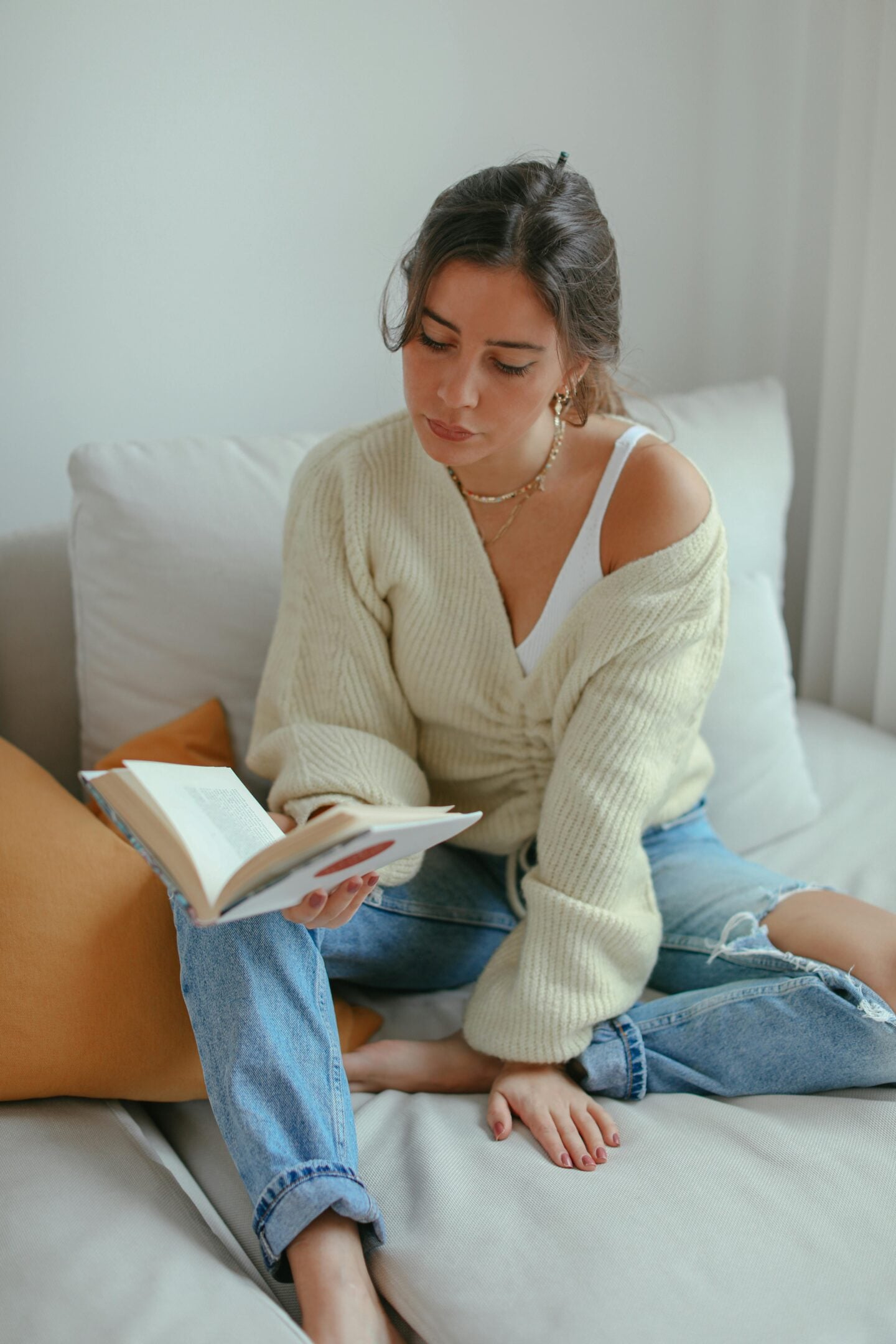 A woman sitting on a sofa indoors, reading a book while wearing a light knit cardigan and jeans, with neutral cushions and soft natural light creating a calm, cozy atmosphere.