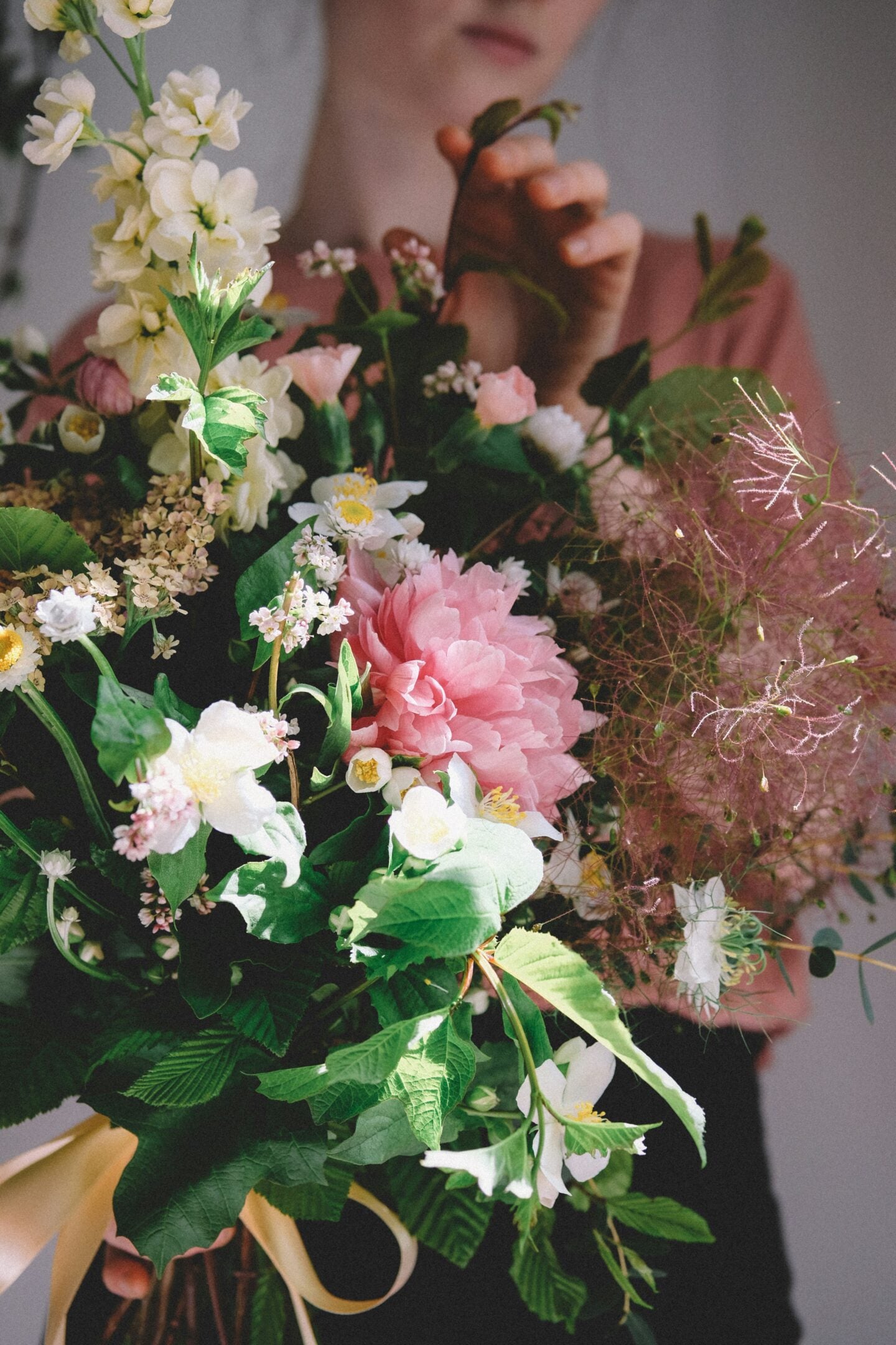 A florist arranging a lush bouquet of mixed flowers, including pink peonies and white blossoms, in soft natural light.