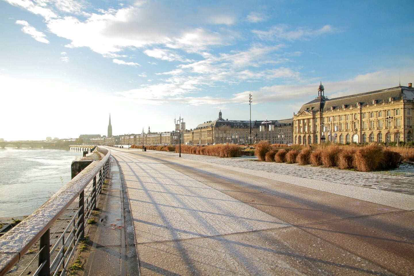 The sunlit promenade along the Garonne River in Bordeaux, lined with historic buildings and winter vegetation beneath a bright sky.