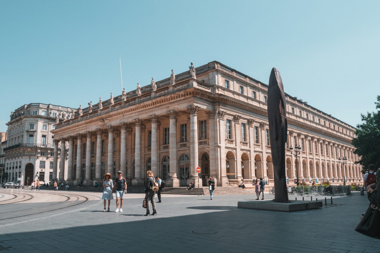 People walk past the Grand Théâtre de Bordeaux, a neoclassical building with tall columns and statues along the roofline, under a clear blue sky.