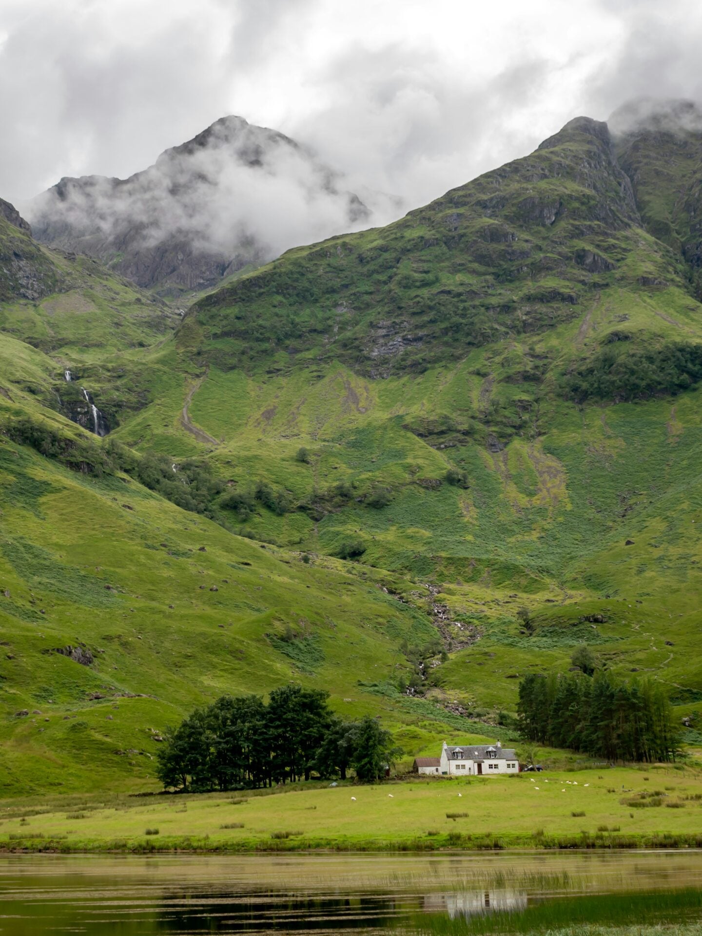 A small white house sits at the base of lush green mountains covered in mist, reflected in a calm lake in the Scottish Highlands.
