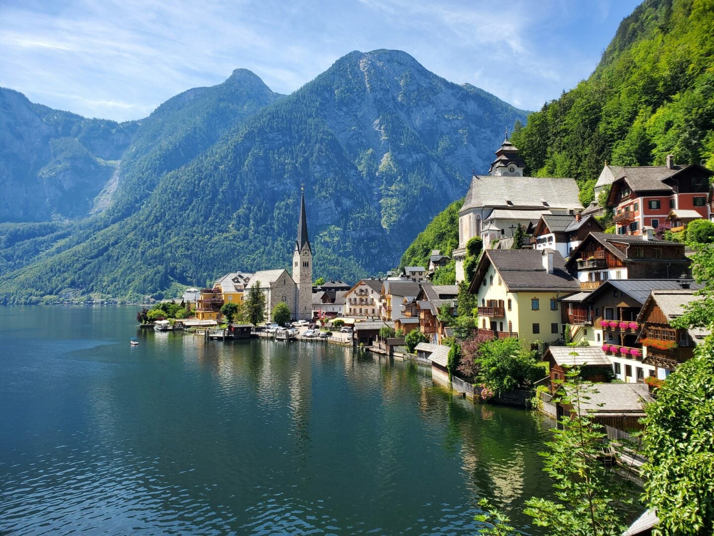 The lakeside village of Hallstatt in Austria, featuring colorful houses, a tall church steeple, and forest-covered mountains reflecting in the calm water on a sunny day.