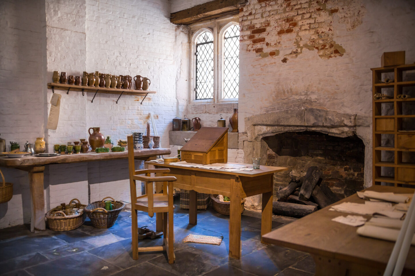 Interior of a historic Tudor kitchen at Hampton Court Palace, showing wooden tables, shelves of pottery, baskets, and a large open fireplace beneath whitewashed and exposed brick walls.