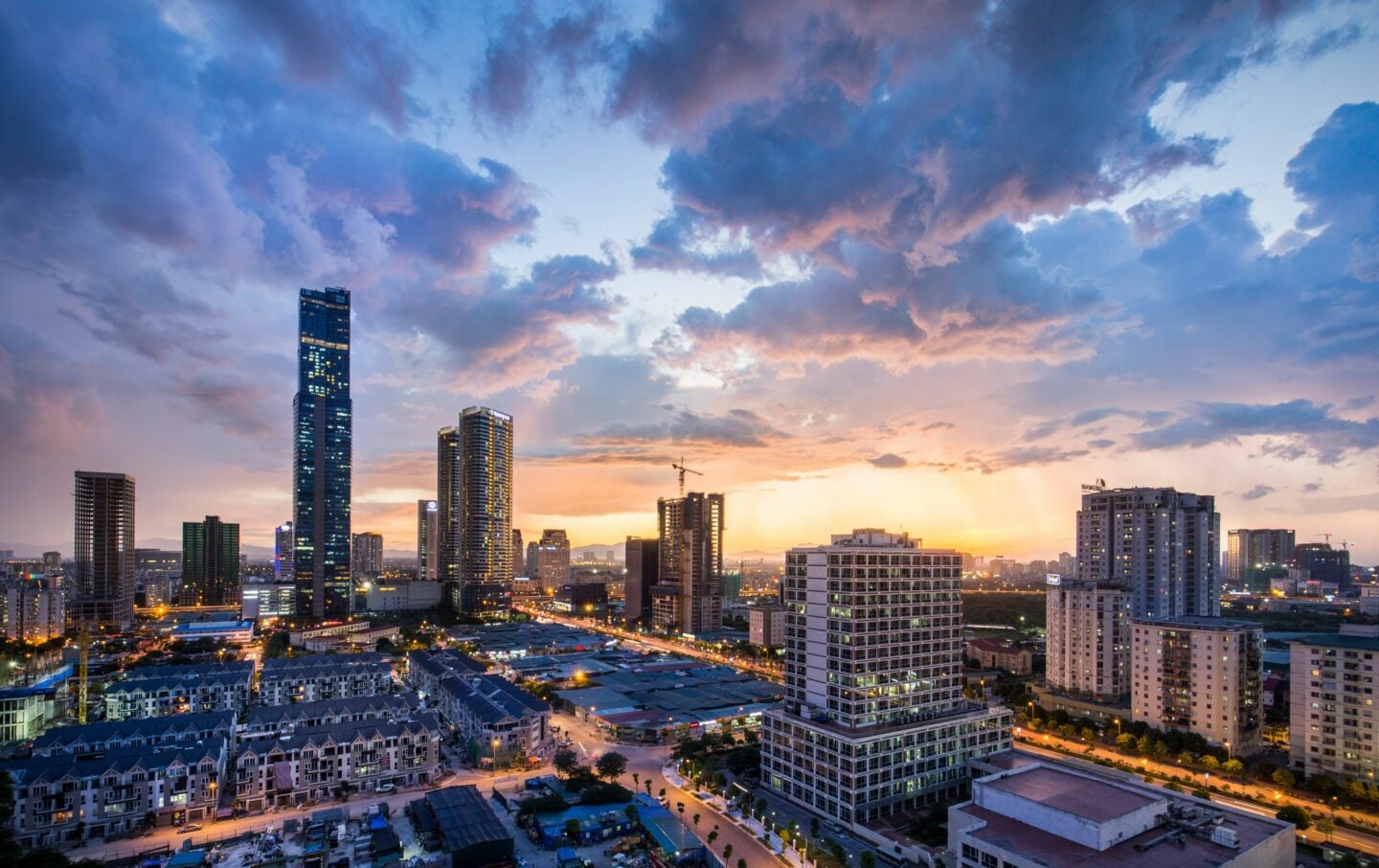 Hanoi, Vietnam city skyline at sunset with modern high-rise buildings glowing under dramatic clouds.