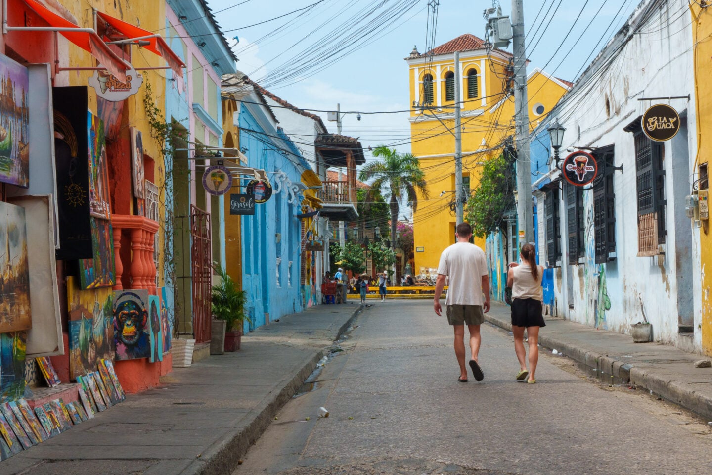 Colorful historic street in Cartagena, Colombia, with bright painted buildings, hanging artwork, shop signs, and pedestrians walking toward a yellow church tower.