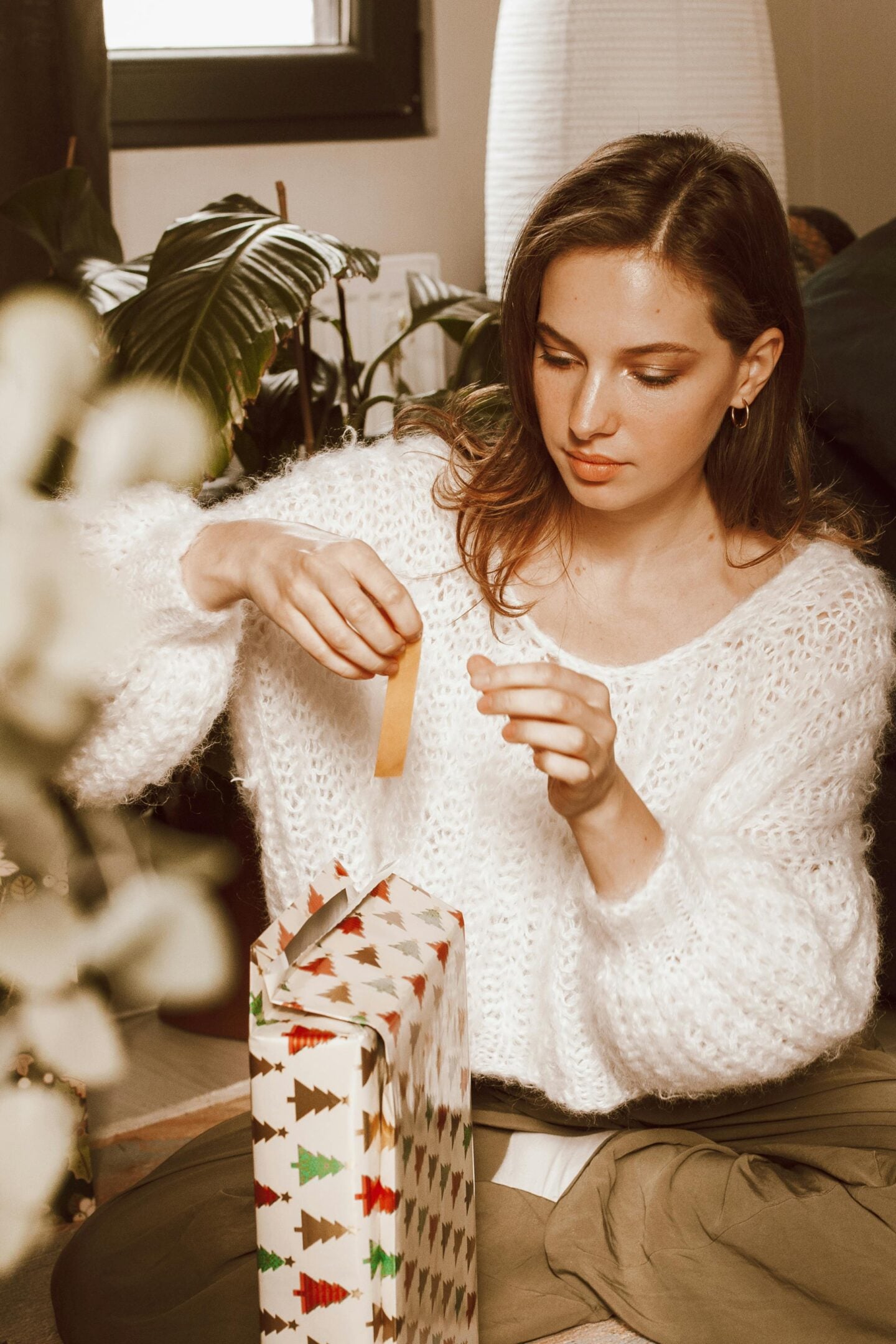 A young woman in a cozy white knitted sweater sits on the floor wrapping a Christmas gift decorated with a tree pattern. She is focused on placing a piece of tape on the present, with soft warm lighting and indoor plants in the background.