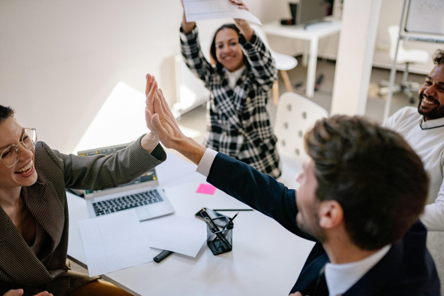 A group of cheerful colleagues in a modern office celebrate a success. Two people in the foreground give each other a high five across a desk scattered with papers and a laptop, while another person stands behind them smiling and raising a document in excitement. A fourth teammate sits nearby laughing, creating an overall atmosphere of teamwork and accomplishment.