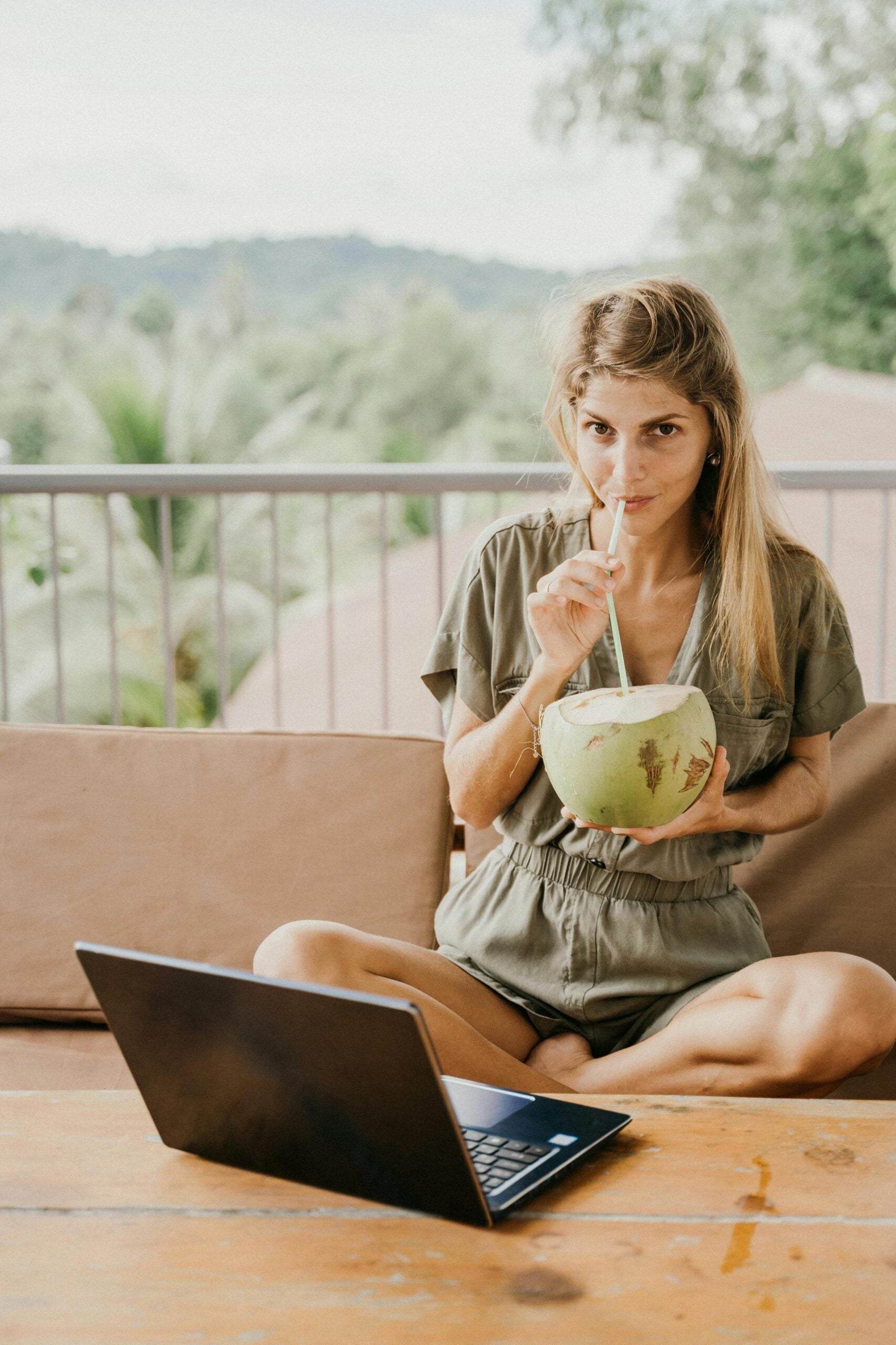 A woman sitting cross-legged on an outdoor sofa, drinking from a fresh coconut while using a laptop on a wooden table, with a lush, green landscape in the background, suggesting a relaxed digital nomad lifestyle.