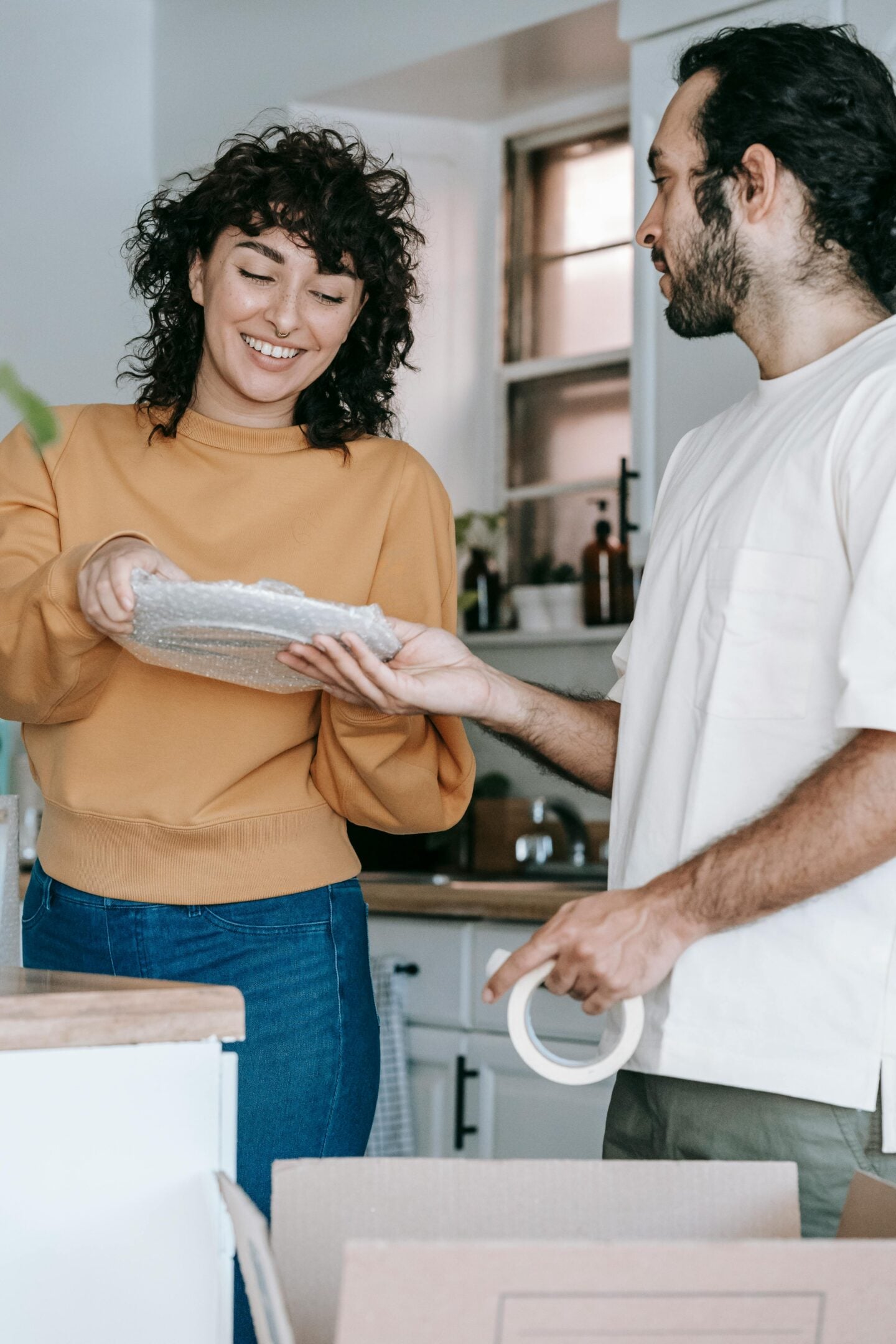 Couple packing and wrapping fragile items with bubble wrap in a kitchen while preparing for a move to a new home.