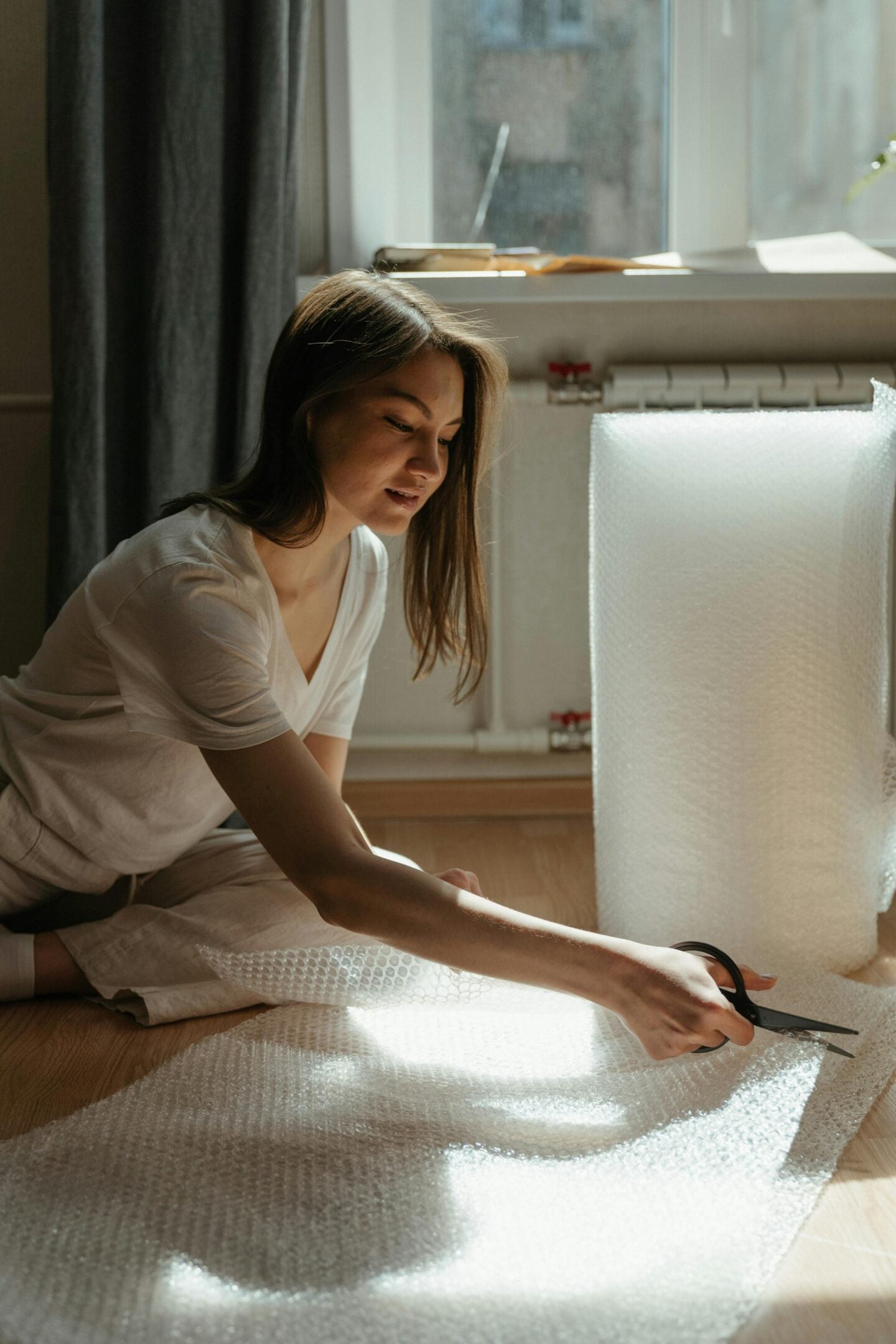 Woman sitting on the floor cutting bubble wrap while carefully packing household items during a home move.