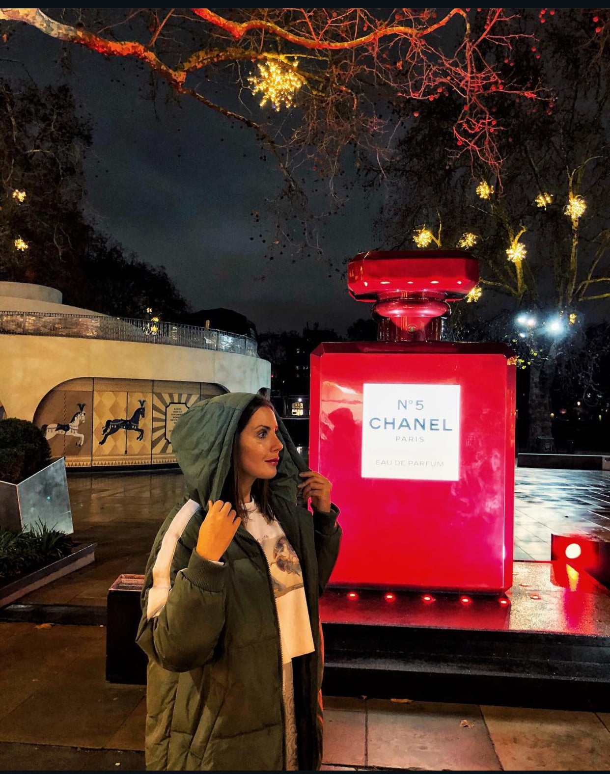 A woman wearing a winter coat standing beside the illuminated Chanel No. 5 Christmas installation in London, surrounded by festive lights during the holiday season.