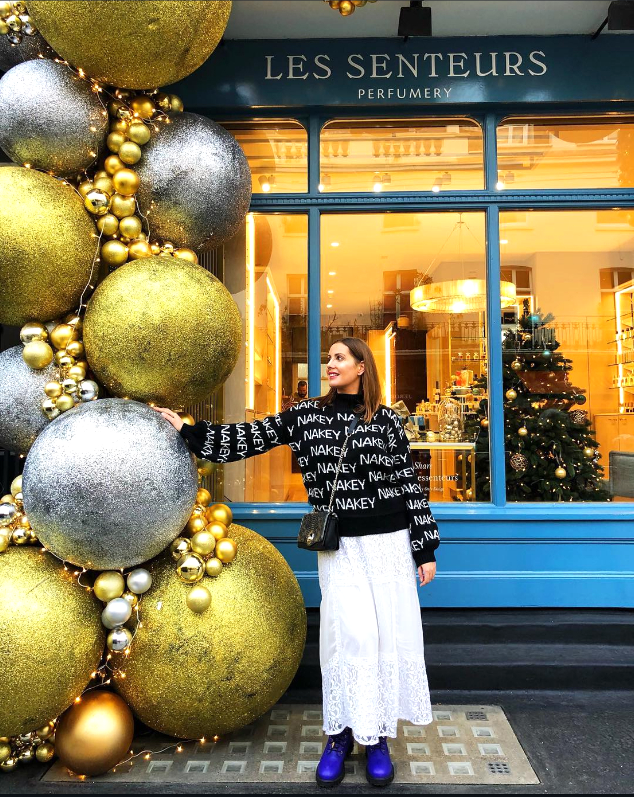 A woman standing in front of Les Senteurs Perfumery in London, surrounded by giant gold and silver Christmas baubles and festive decorations during the winter season.