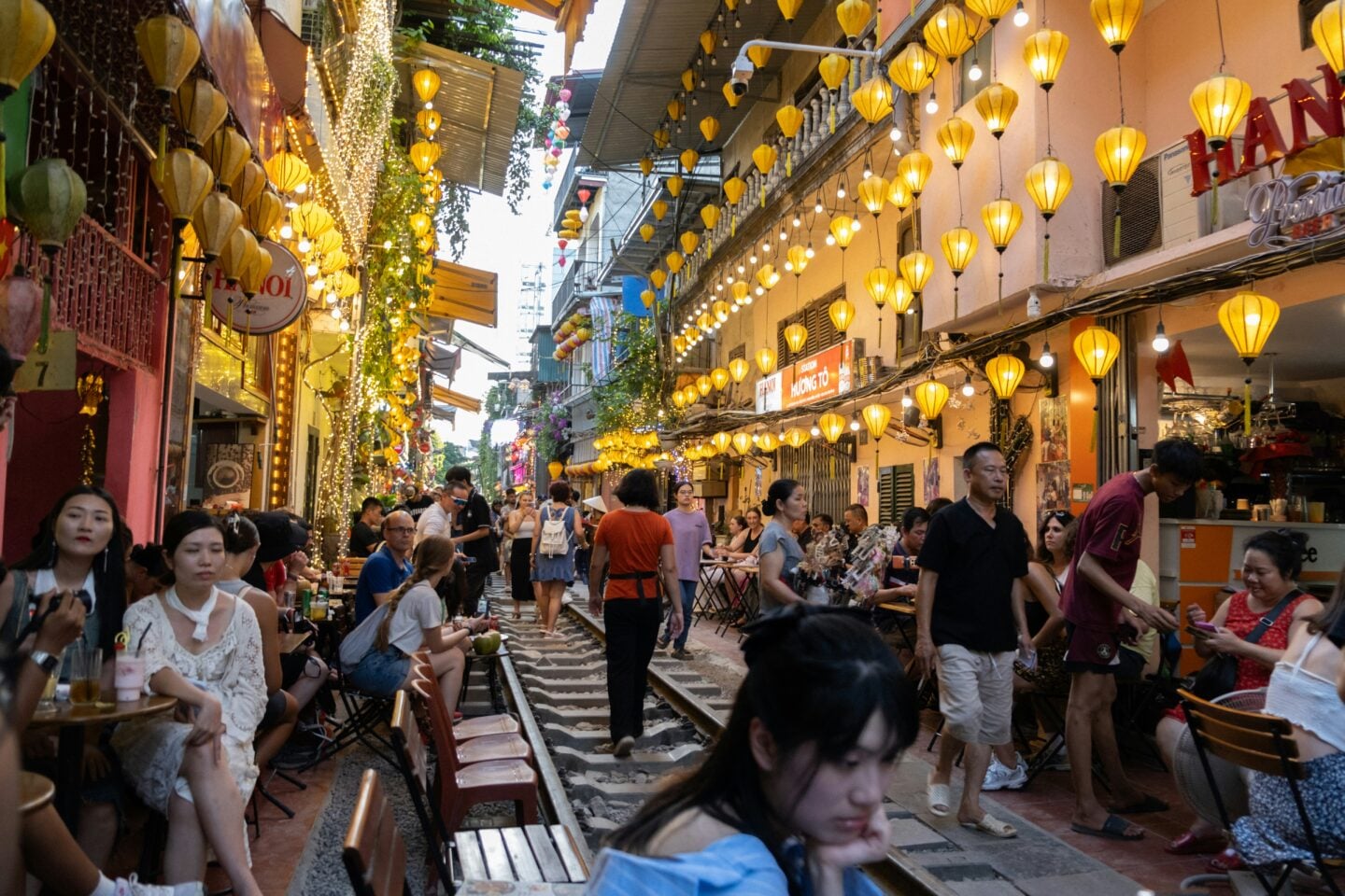 People walking and dining along the busy Train Street in Hanoi, Vietnam, illuminated by hanging lanterns and street lights in the evening.