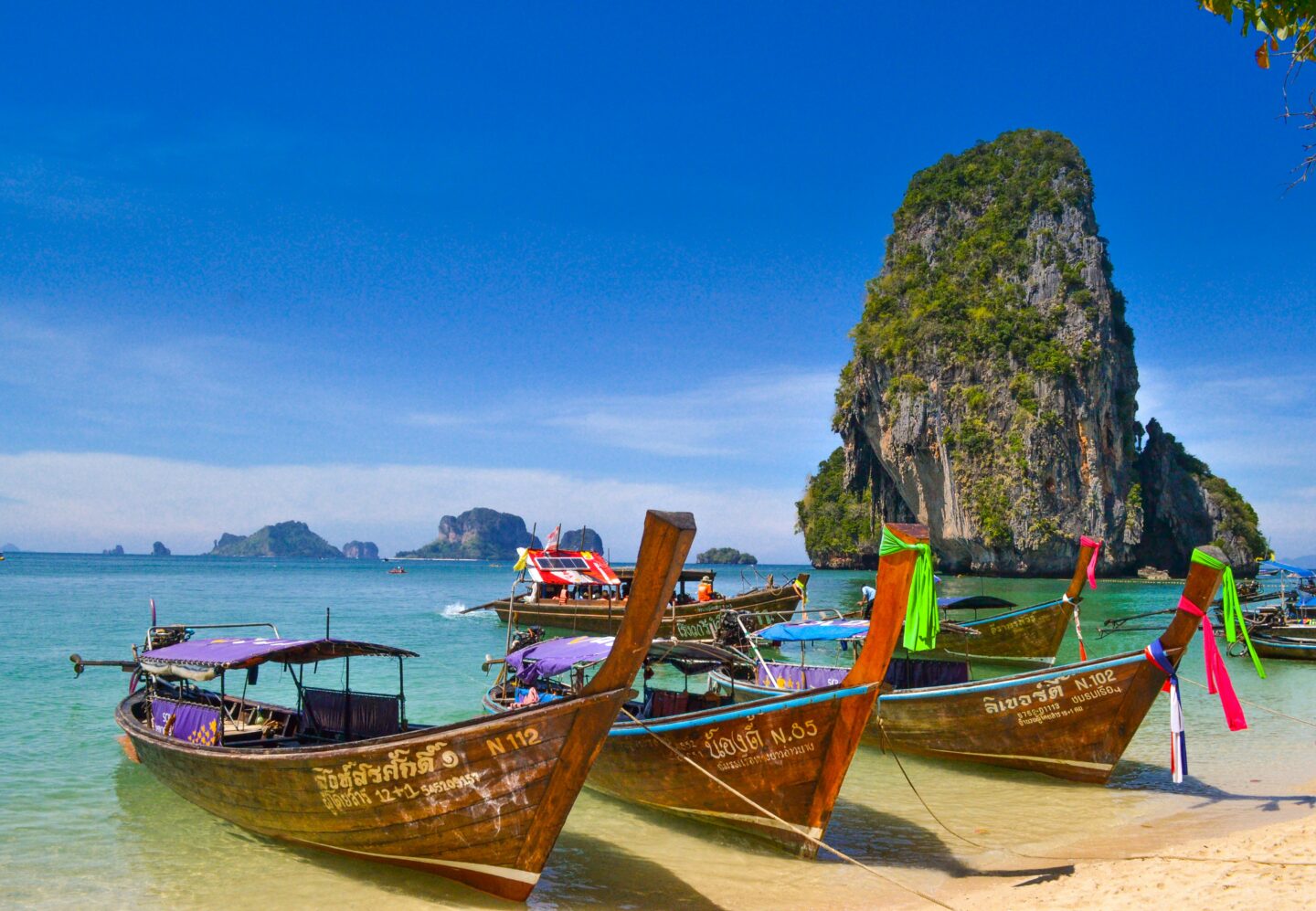 Traditional long-tail boats anchored in turquoise water at Phra Nang Beach in Krabi, Thailand, with dramatic limestone cliffs in the background.