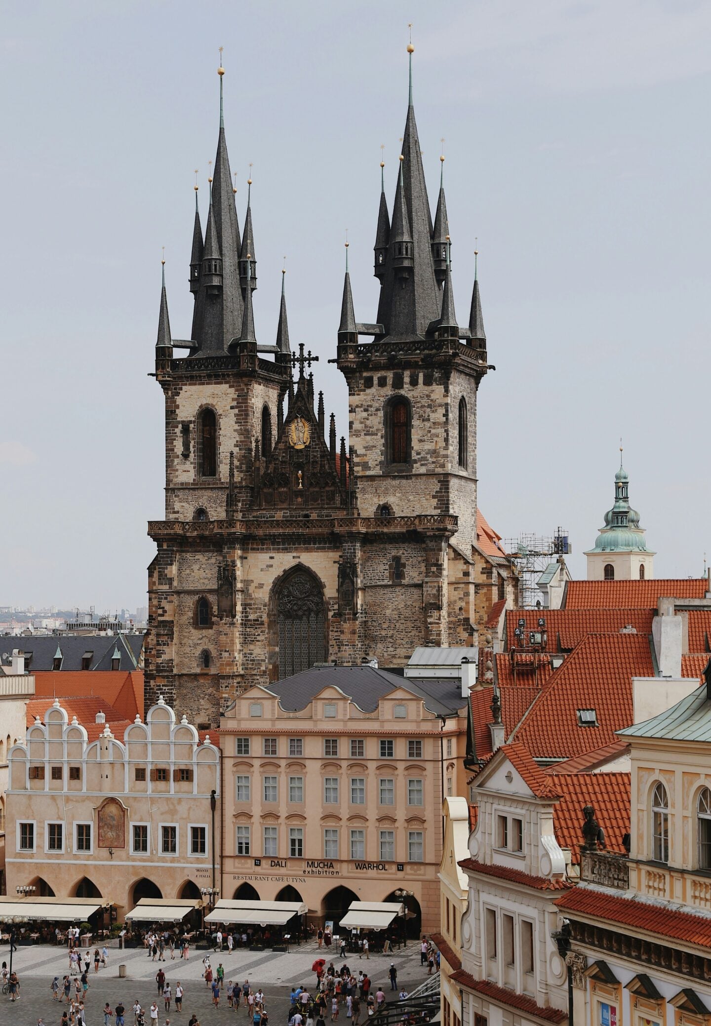 The Church of Our Lady before Týn with its tall Gothic spires towering over Old Town Square in Prague, Czechia, with historic pastel buildings and crowds in the square below.