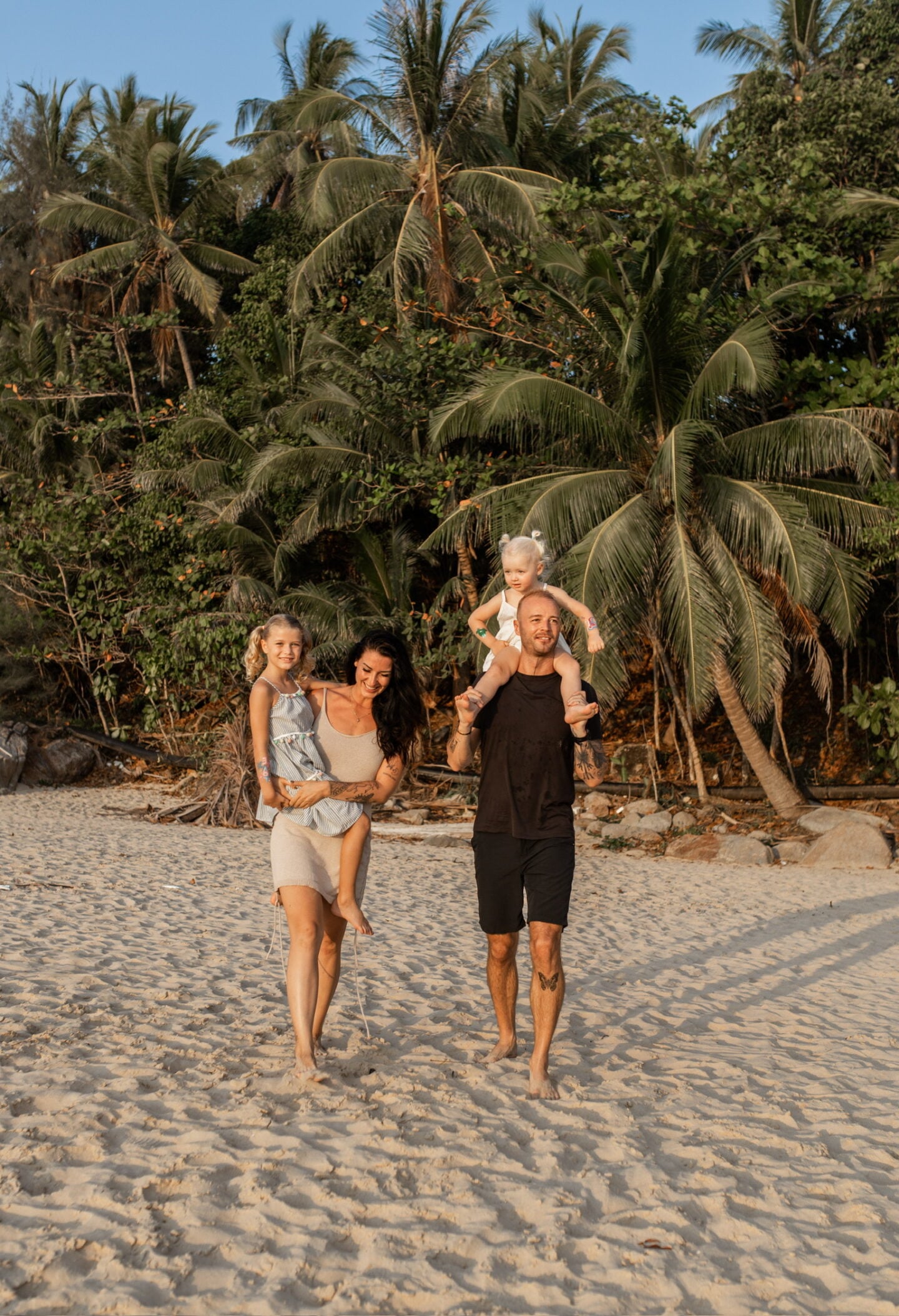 Family walking barefoot along a tropical beach in Thailand lined with palm trees, enjoying a relaxed moment by the sea.