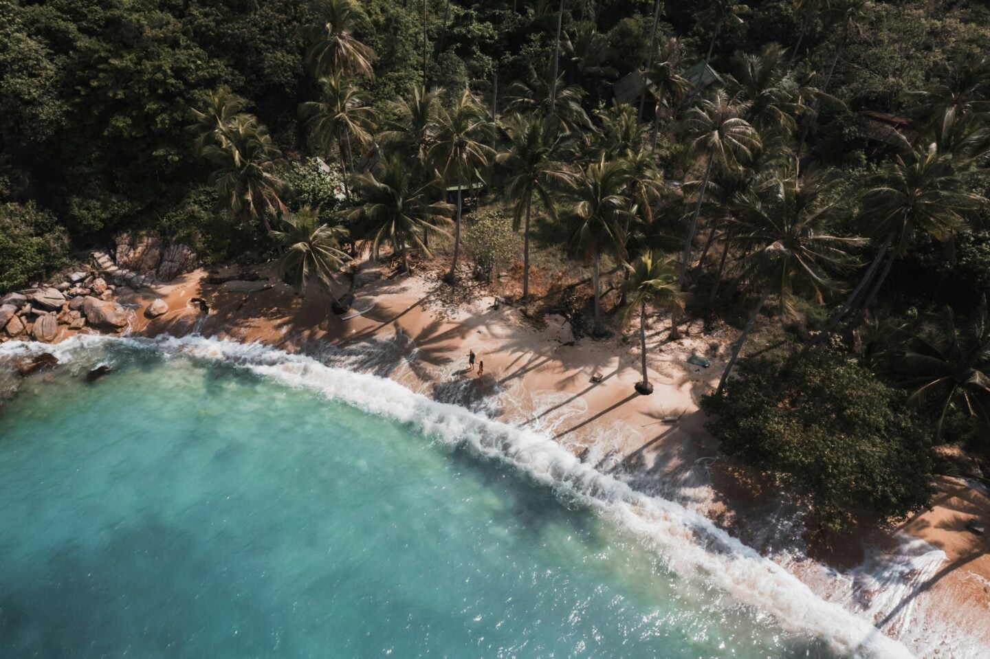 Aerial view of a quiet tropical beach in Thailand, with turquoise water, palm trees, and waves gently washing onto the sandy shore.