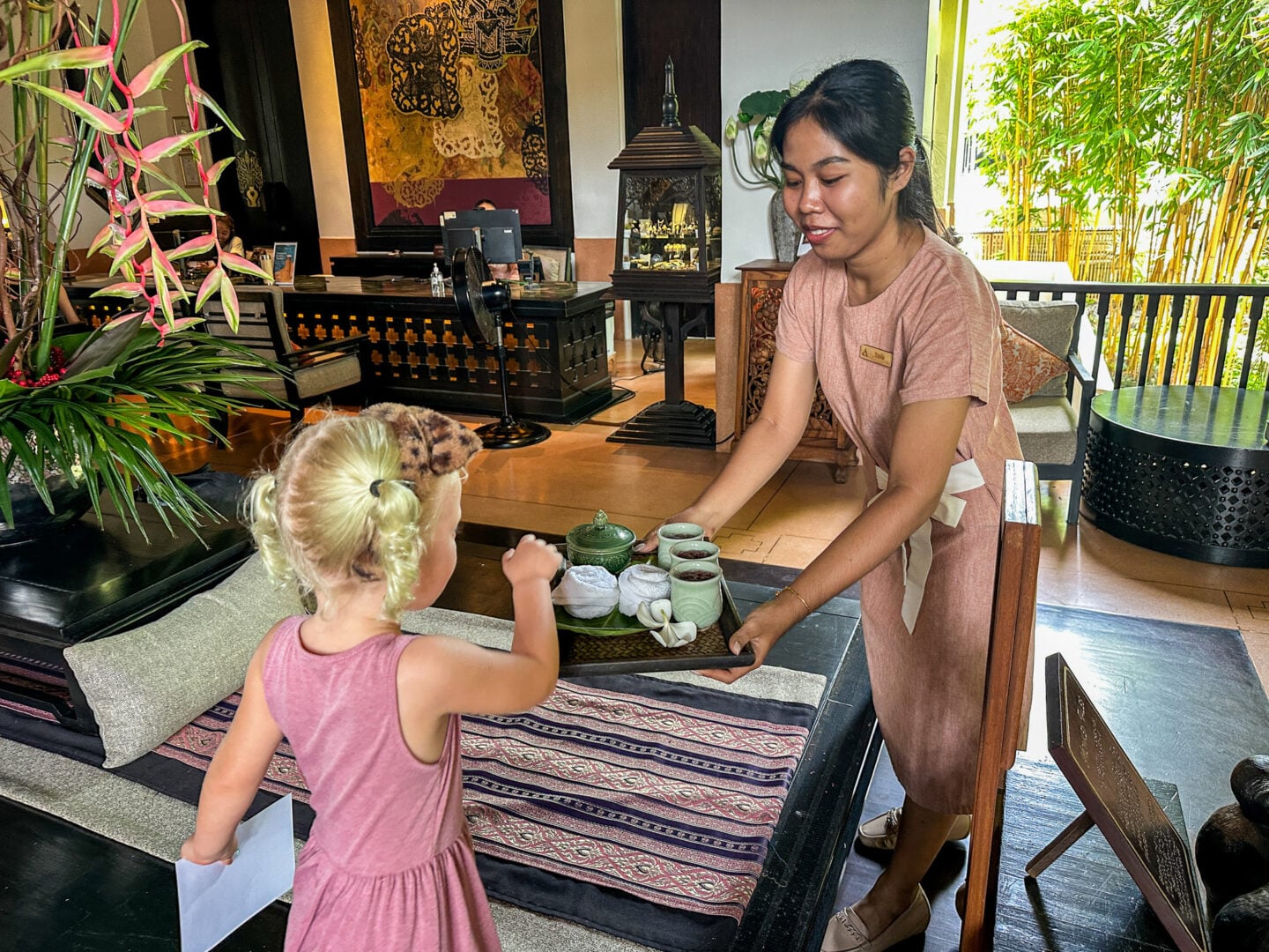 Hotel staff member welcoming a young child with a tray of drinks in a Thai resort lobby decorated with plants and wooden furnishings.