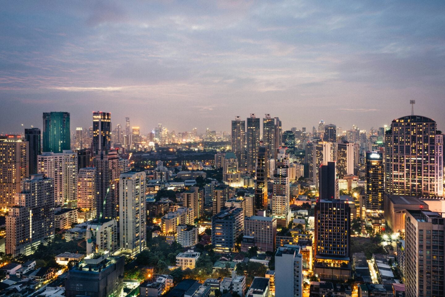 Evening city skyline of Bangkok with illuminated high-rise buildings and dense urban landscape at dusk.