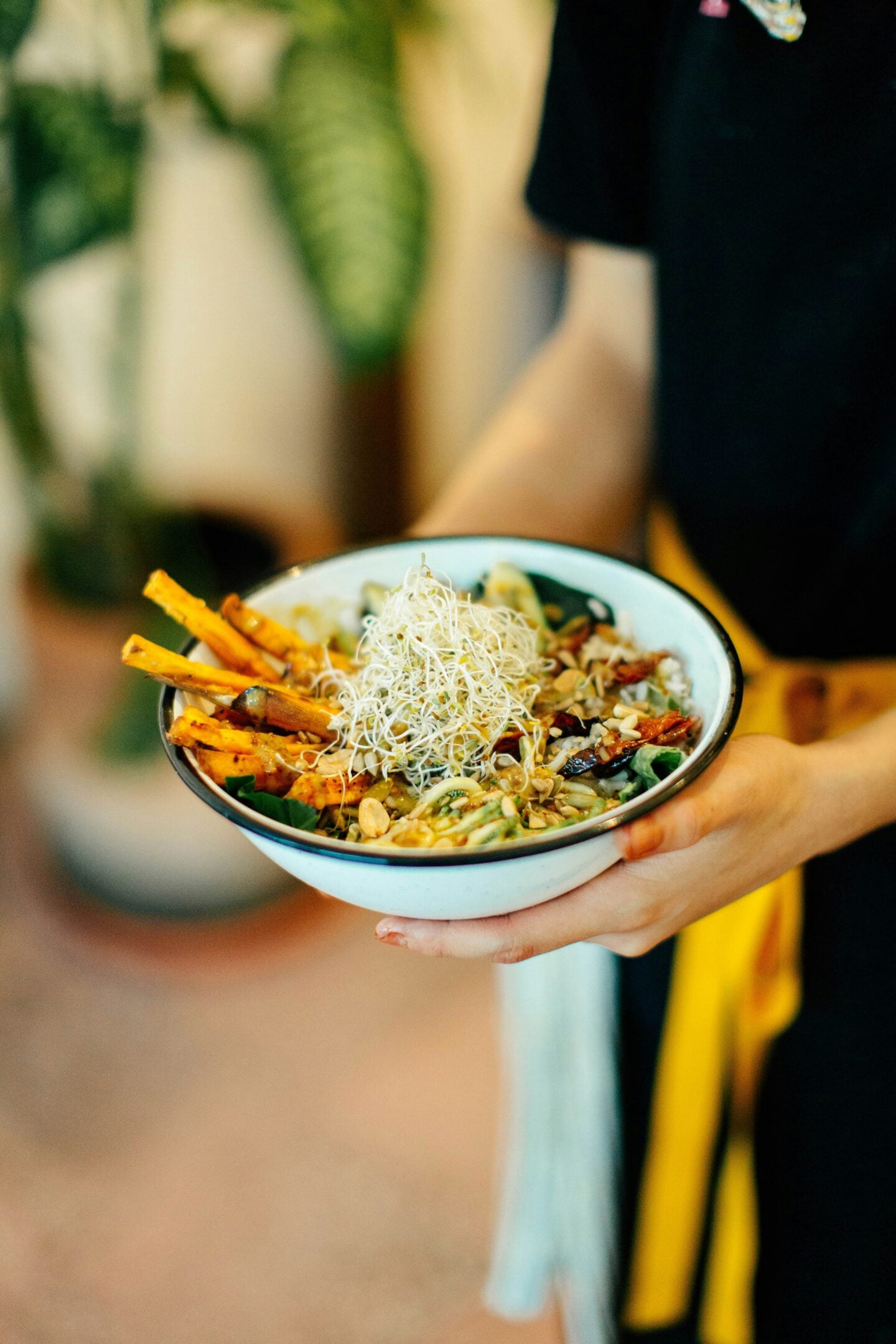 Close-up of a bowl of colorful plant-based food topped with sprouts and seeds, held by a person indoors.