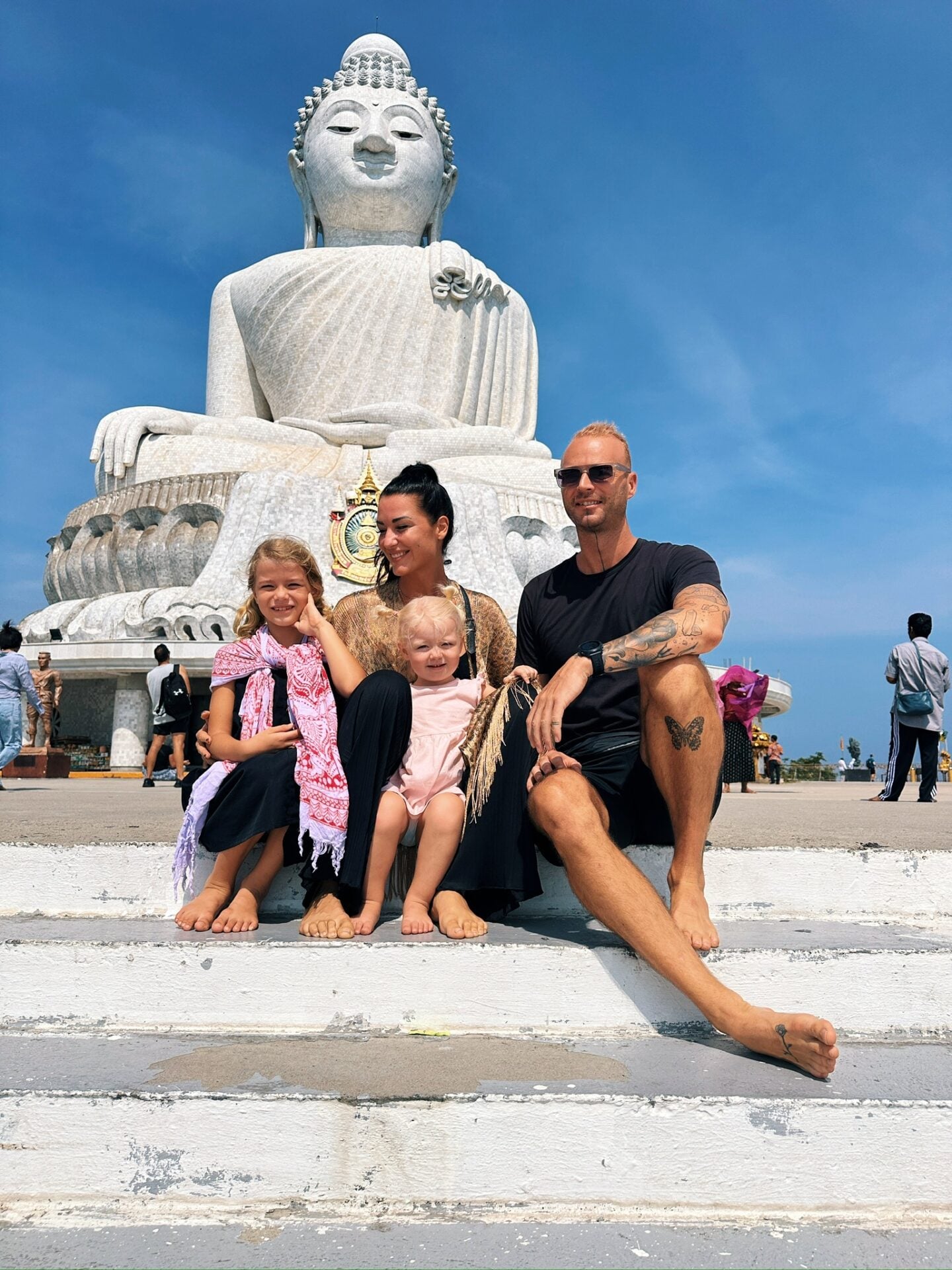 Family sitting on steps in front of the Big Buddha statue in Phuket, Thailand, with the white statue rising behind them under a clear blue sky.