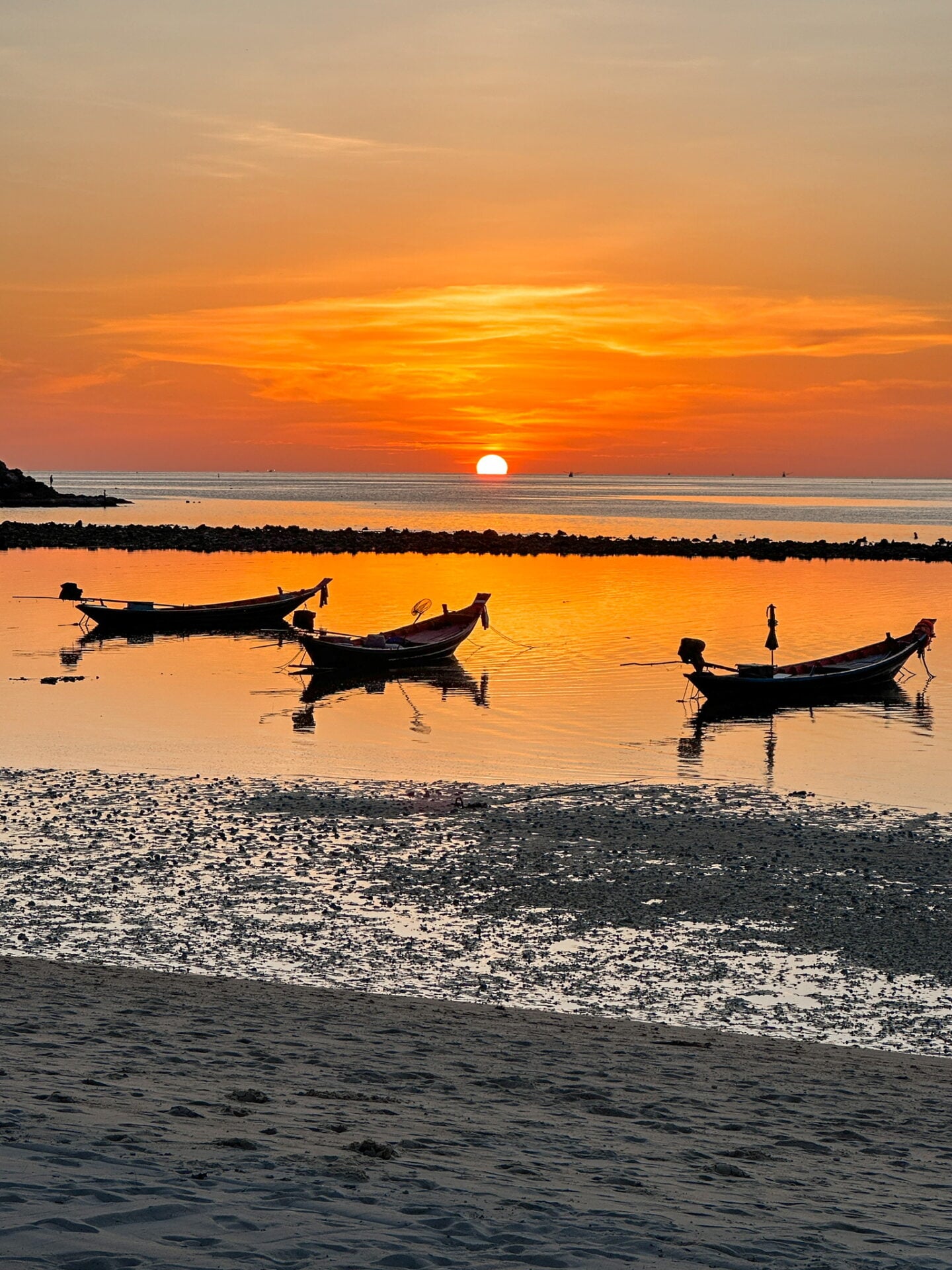Long-tail boats floating on calm water at sunset, silhouetted against an orange sky along a Thai beach.