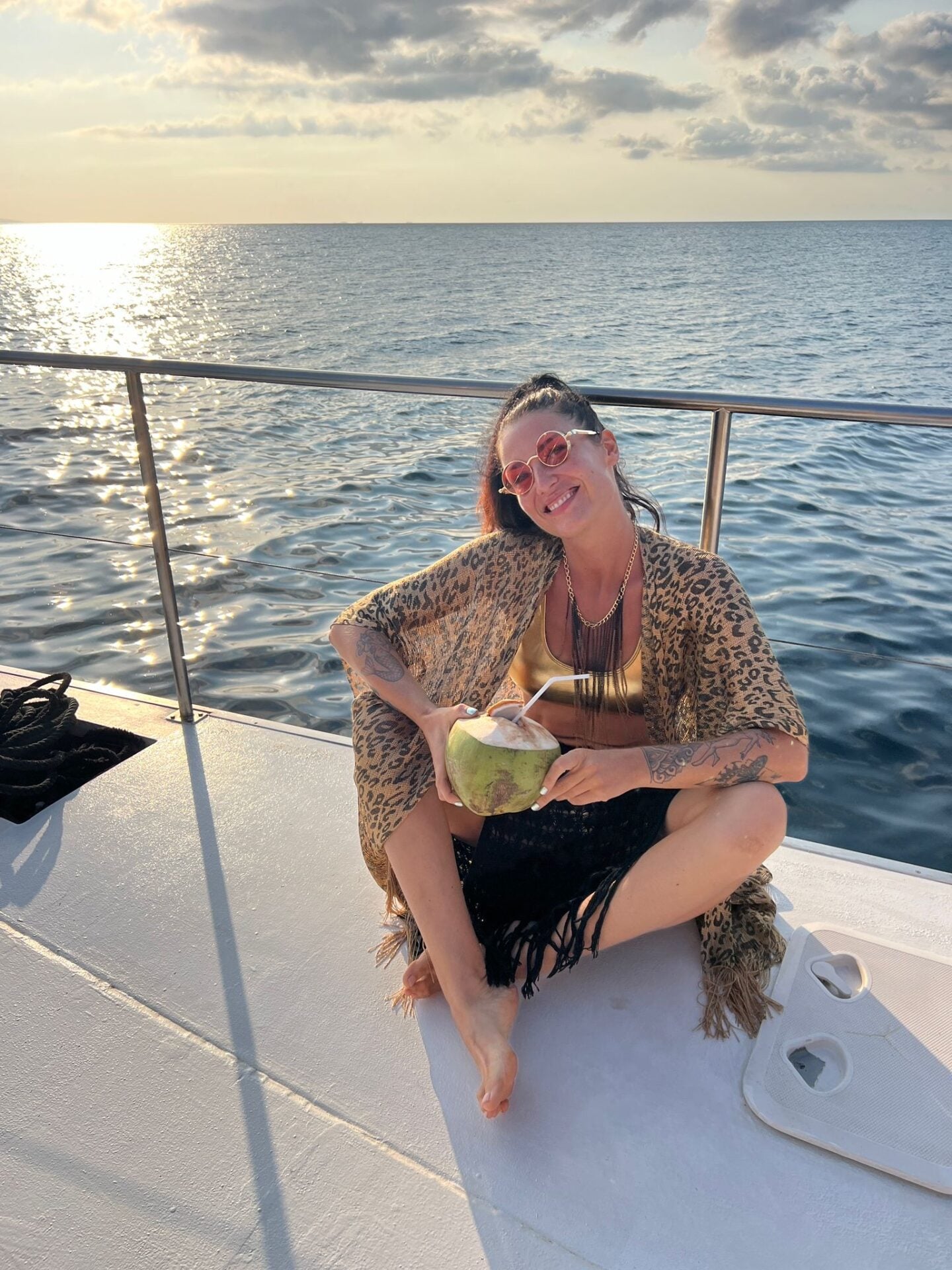 Woman sitting on a boat deck holding a fresh coconut drink, with open sea and sunlight reflecting on the water behind her.