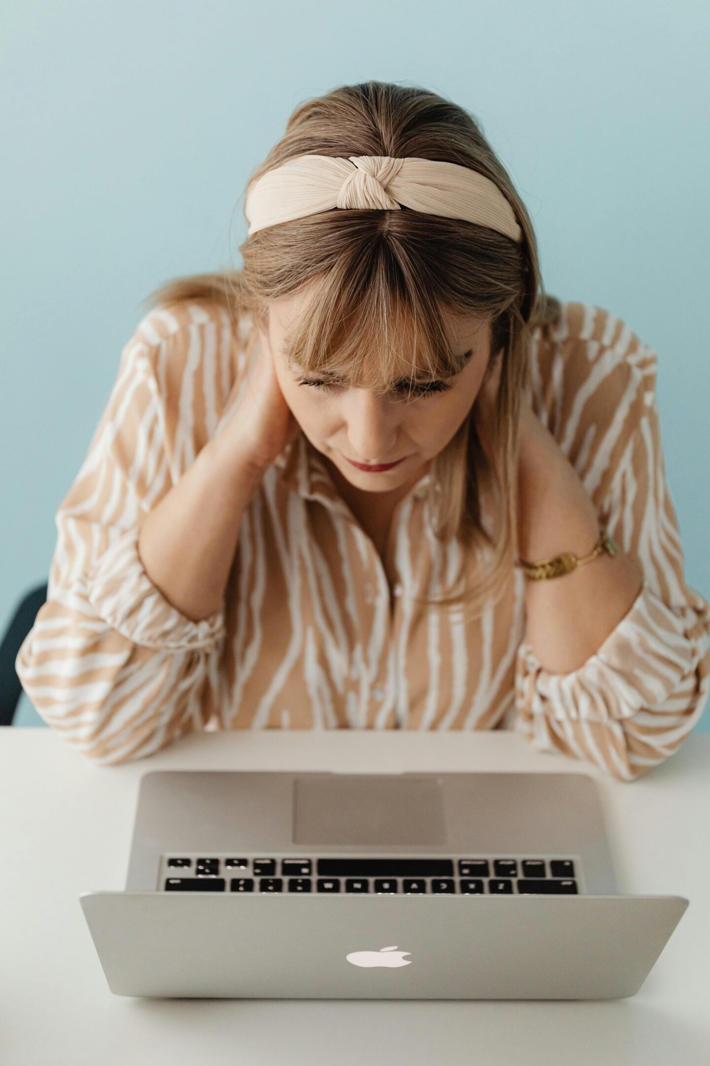 A woman wearing a beige headband and patterned blouse looks down at her laptop with her hands on her neck, appearing focused.