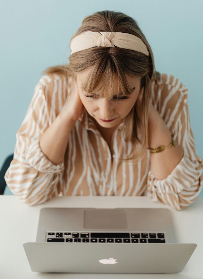 A woman wearing a beige headband and patterned blouse looks down at her laptop with her hands on her neck, appearing focused.