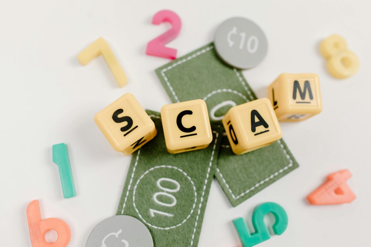 Yellow letter blocks spelling “SCAM” placed on top of felt play money, surrounded by colorful plastic numbers.