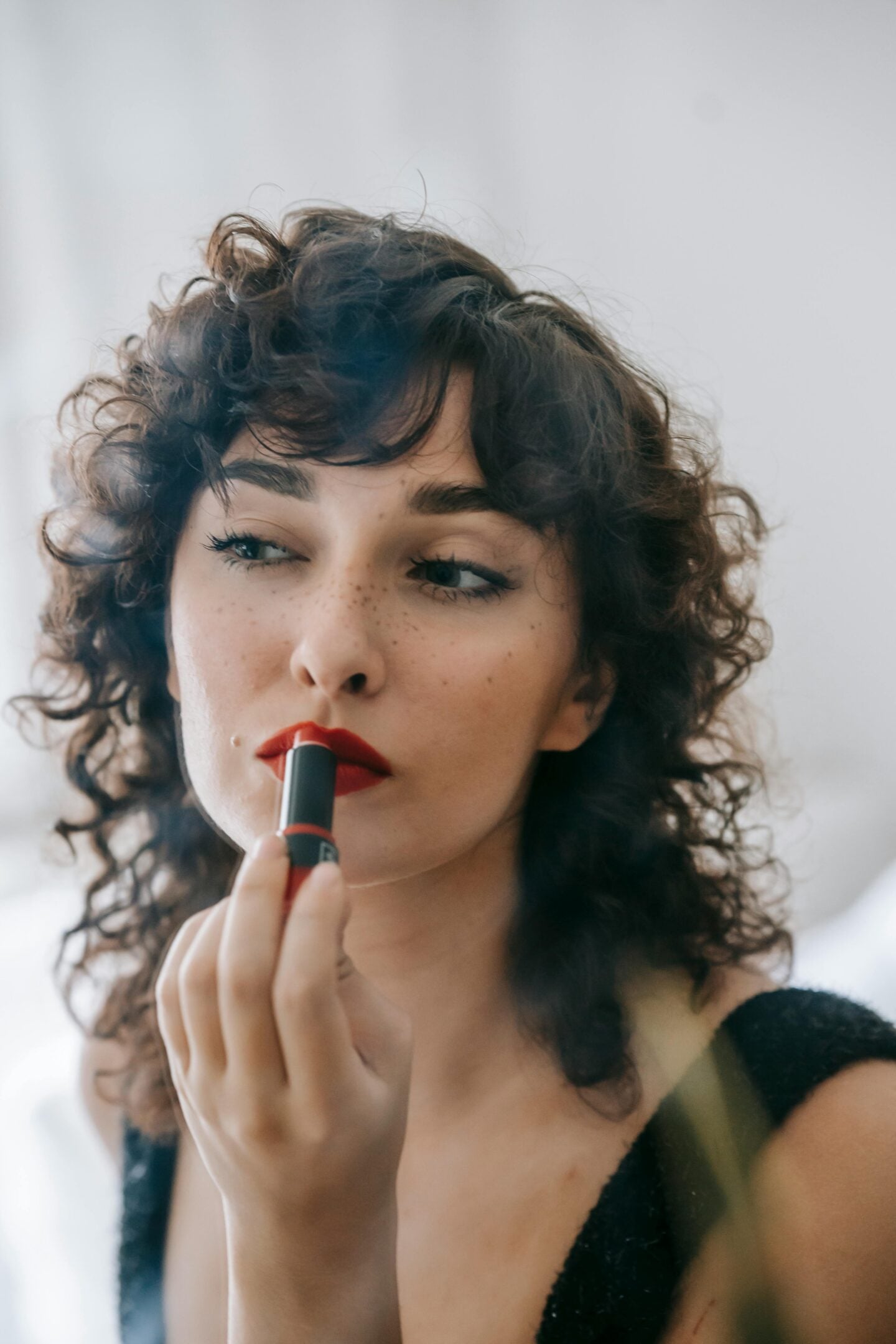 A woman applying red lipstick while looking into a mirror, with curly hair and natural freckles.