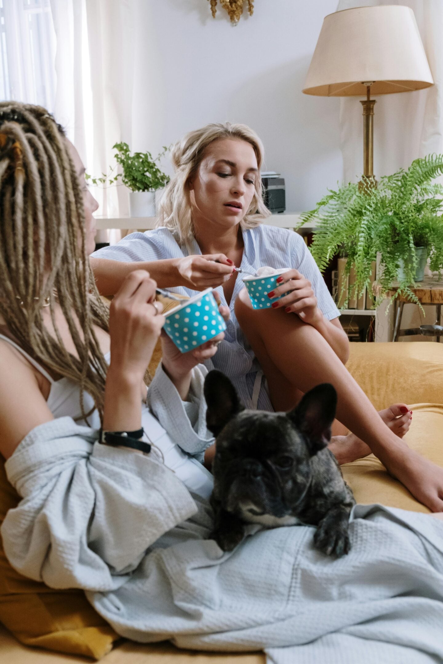 Two women lounging at home eating ice cream, with a French bulldog resting on a blanket beside them.
