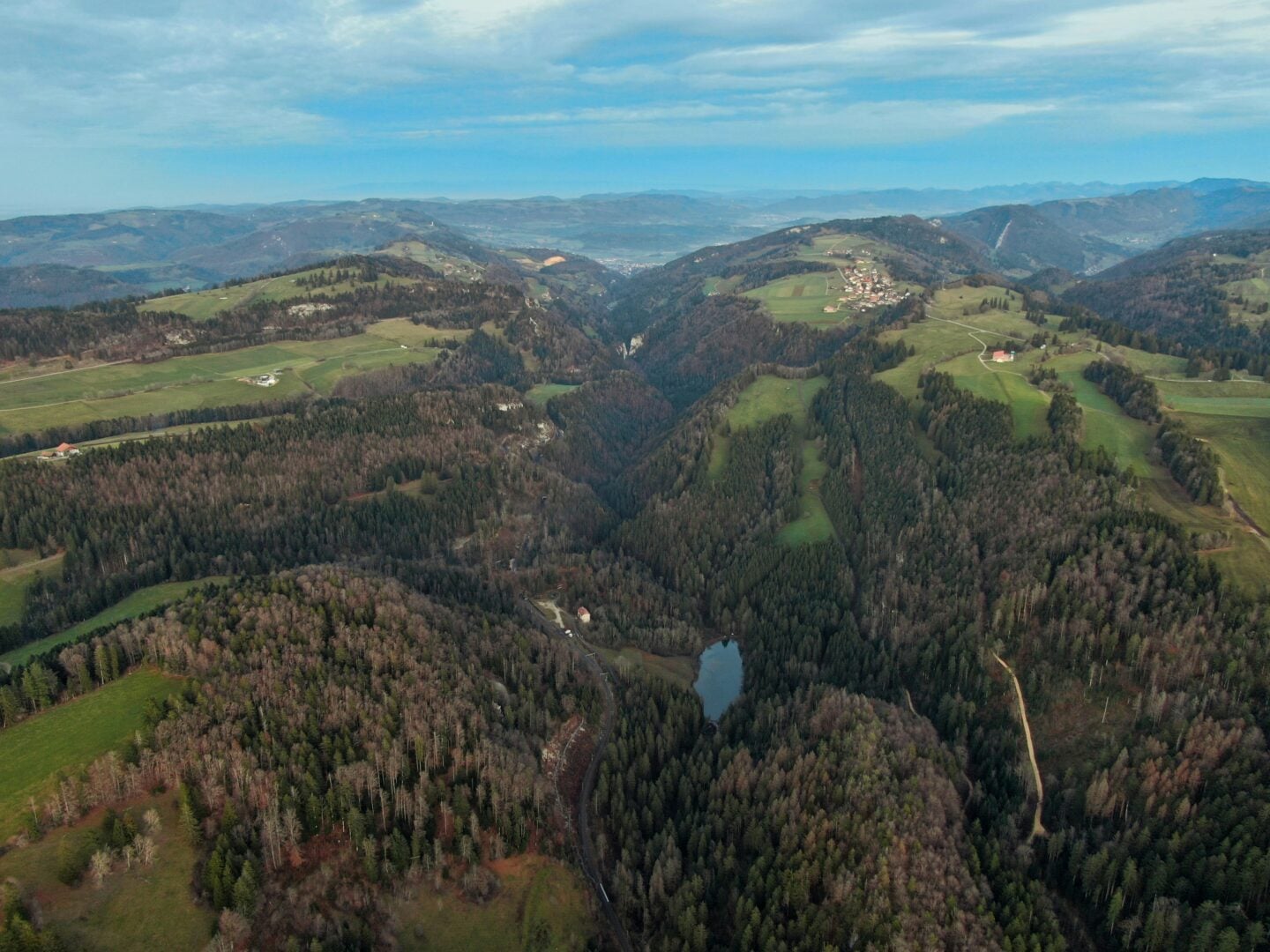 Aerial view of the Swiss Jura landscape featuring rolling hills, dense forests, deep valleys, and scattered villages under a soft blue sky.