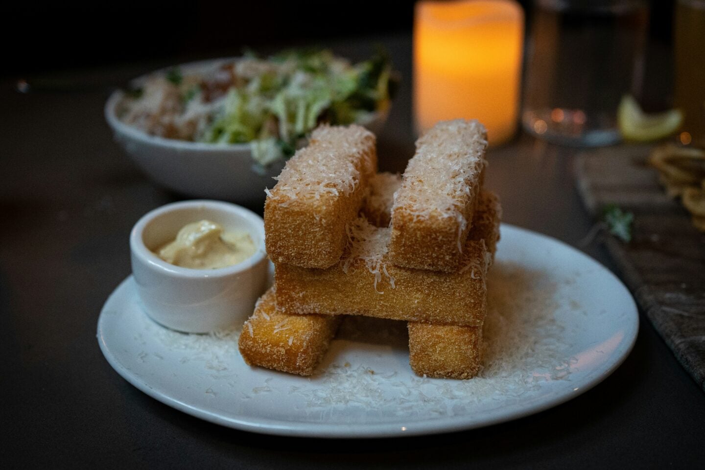 A plate of crispy polenta fries stacked neatly and topped with grated cheese, served with a small dish of creamy dipping sauce in a dimly lit Seattle restaurant.