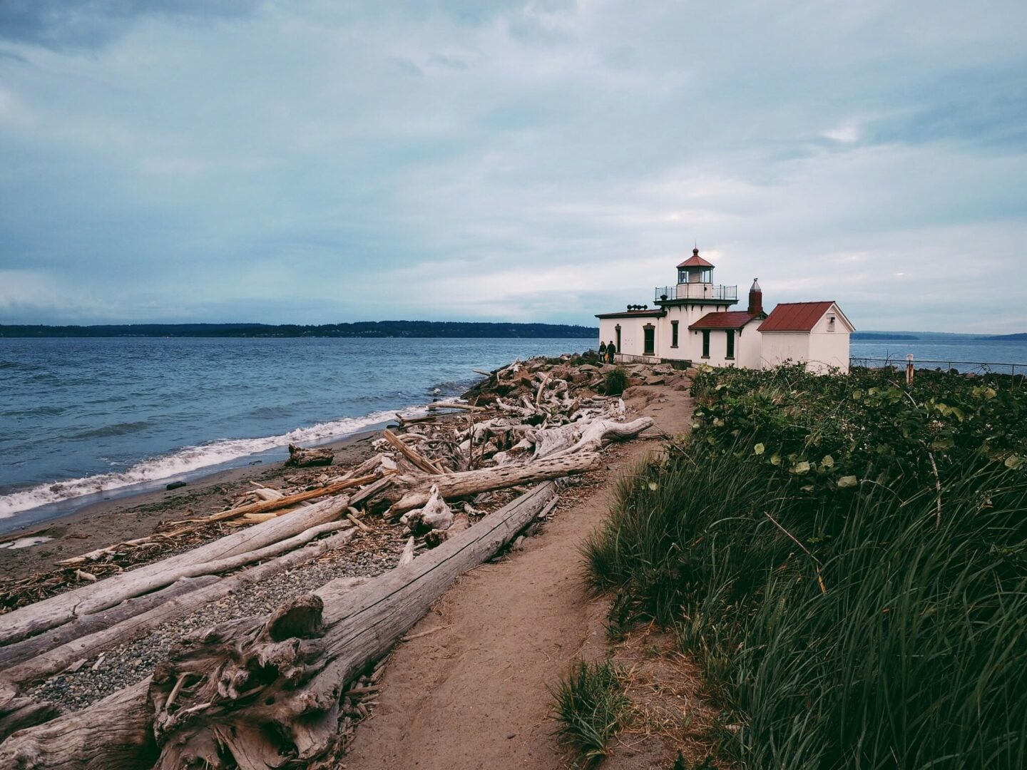The West Point Lighthouse at Discovery Park sits on a driftwood-covered beach with waves and cloudy skies along the Seattle shoreline.