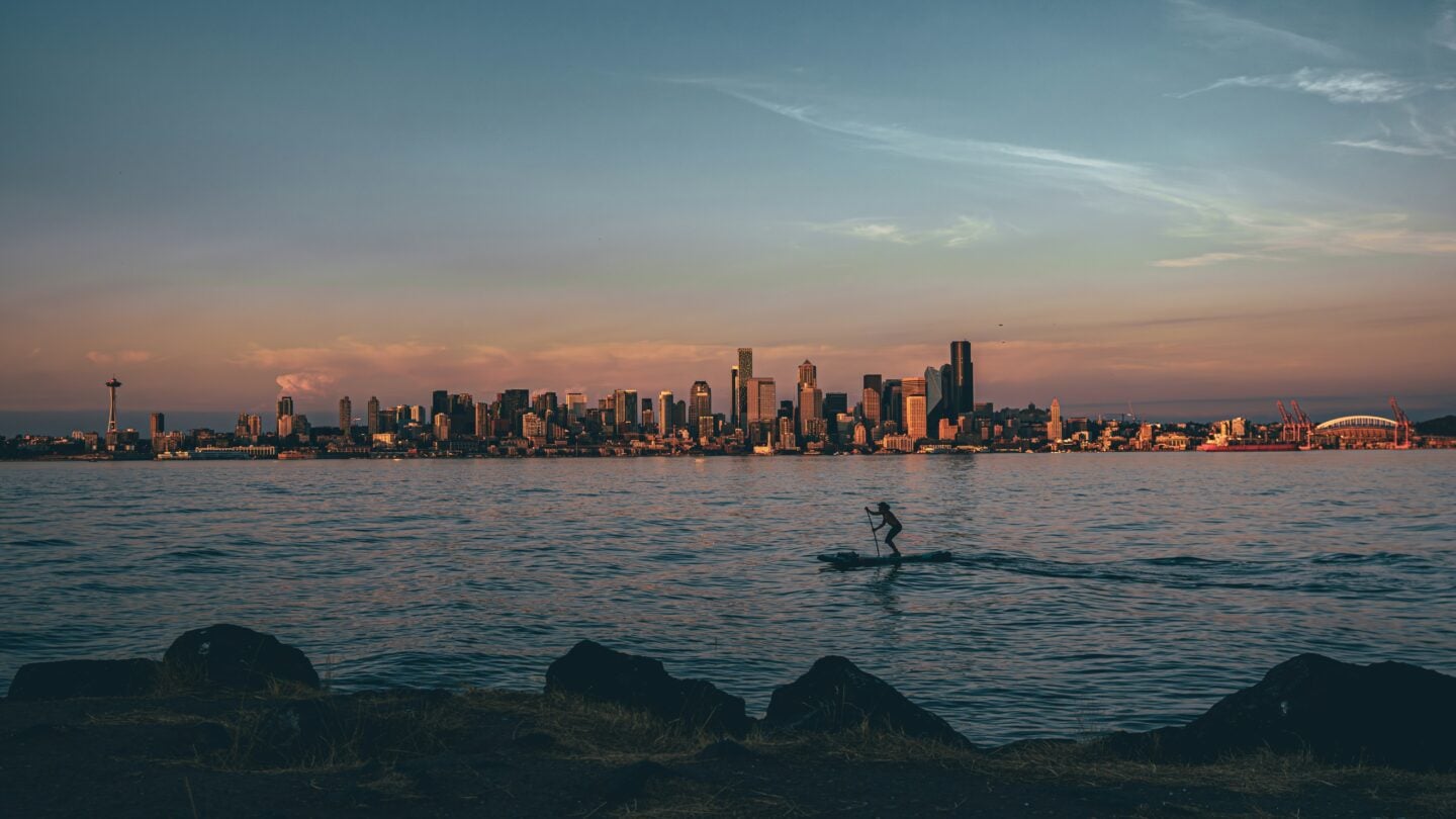 The Seattle skyline glows in golden sunset light as a paddleboarder moves across the water near Alki Beach.